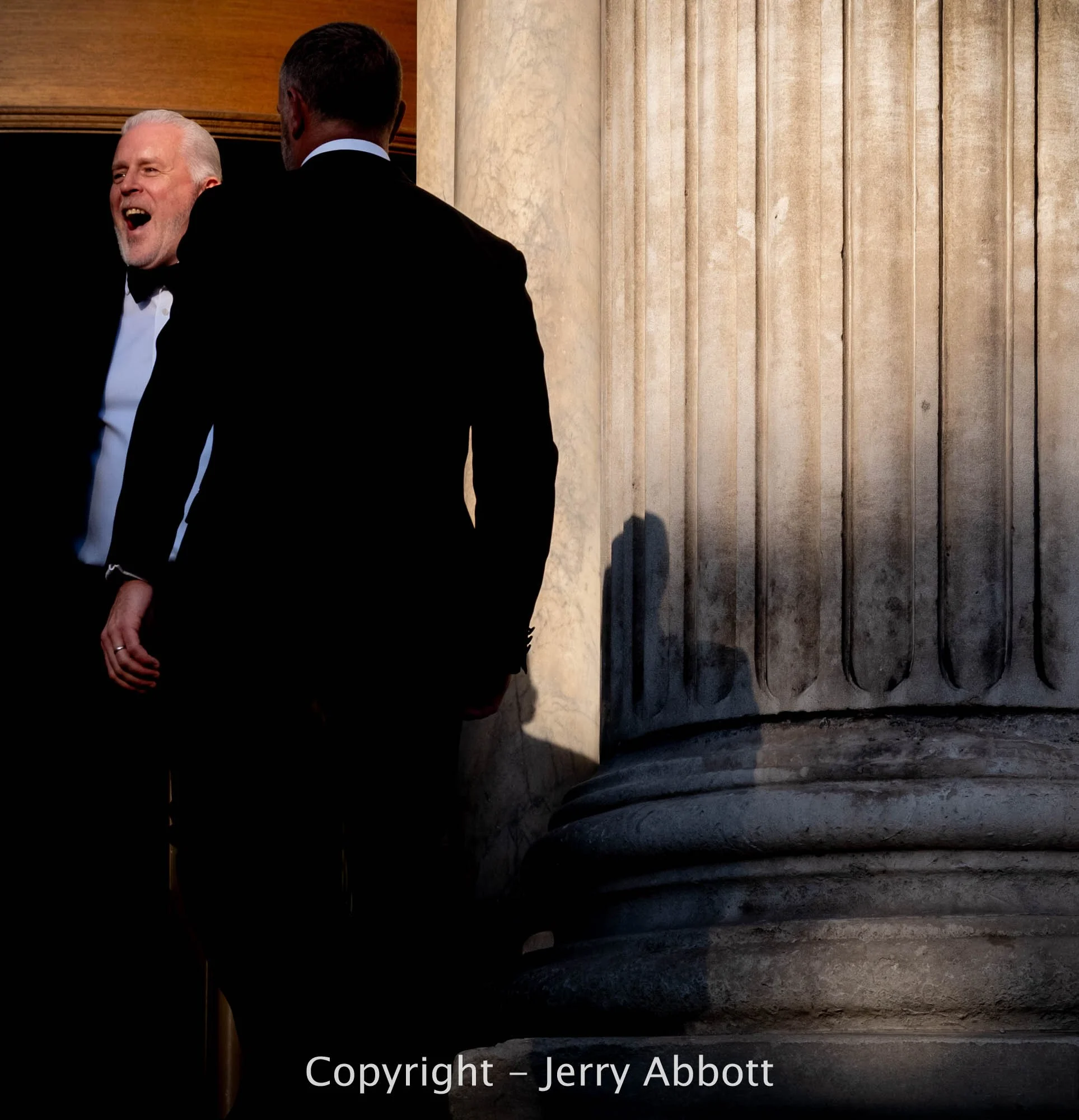 Two men in black-tie attire stand in the doorway of St Paul's Cathedral, framed by dramatic light and shadow beside a large stone column.