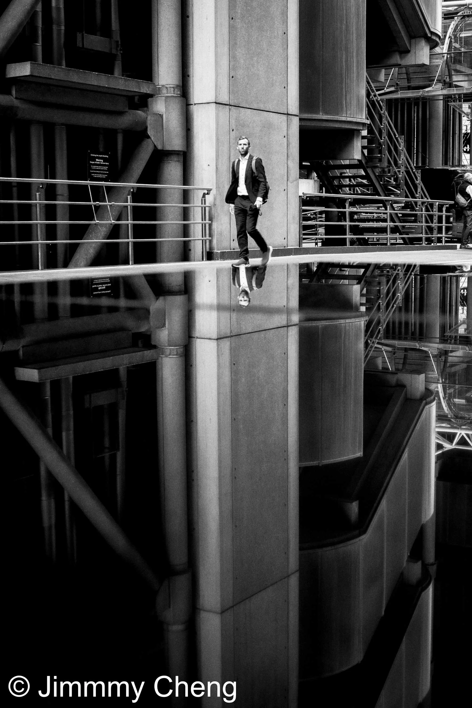Black and white London street photography showing a man walking through modern architecture, with strong geometric lines and his reflection mirrored across a reflective surface.