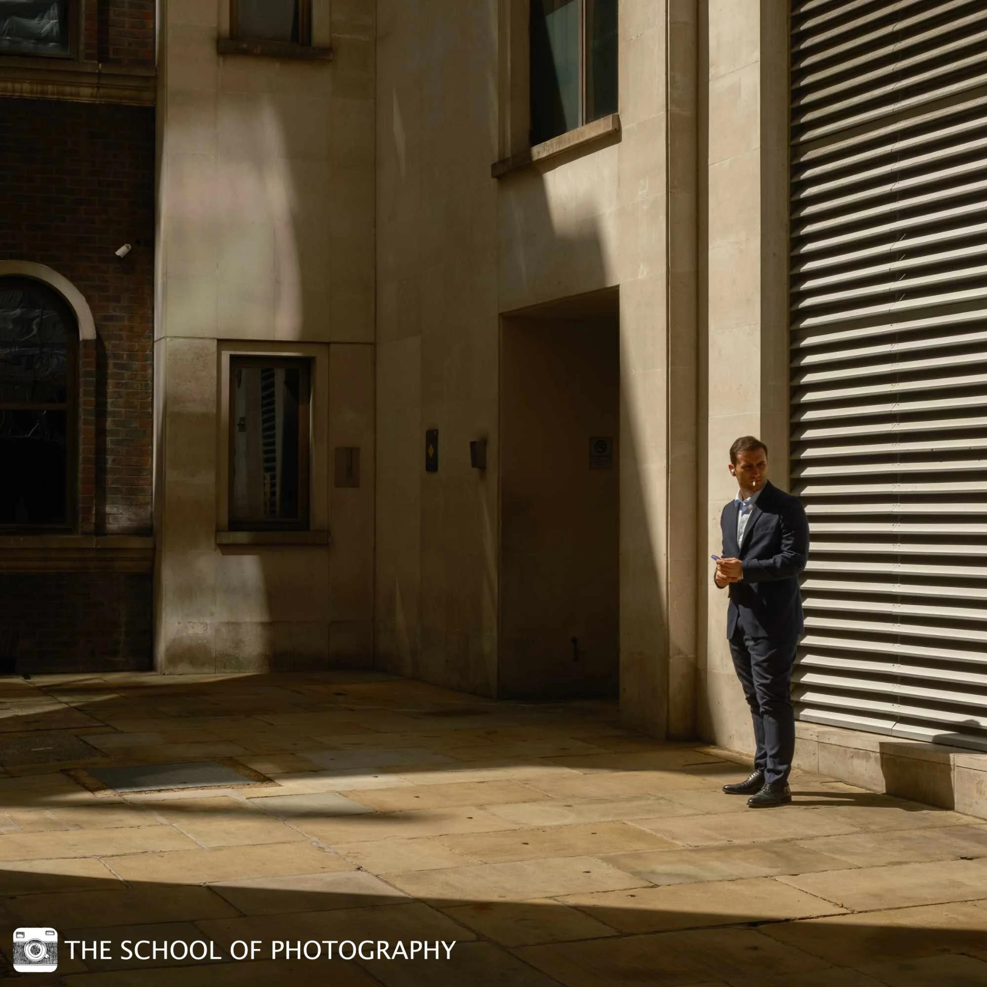 Colour street photography image of a man in a suit standing in harsh sunlight against an urban building, framed by strong architectural lines and deep shadows.
