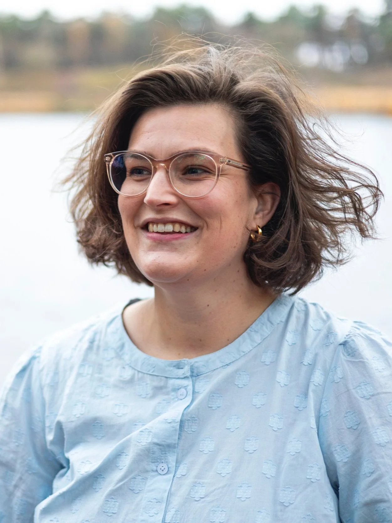 Persoon met bril en blauw blouse staat buiten met een wazige achtergrond van een meer.