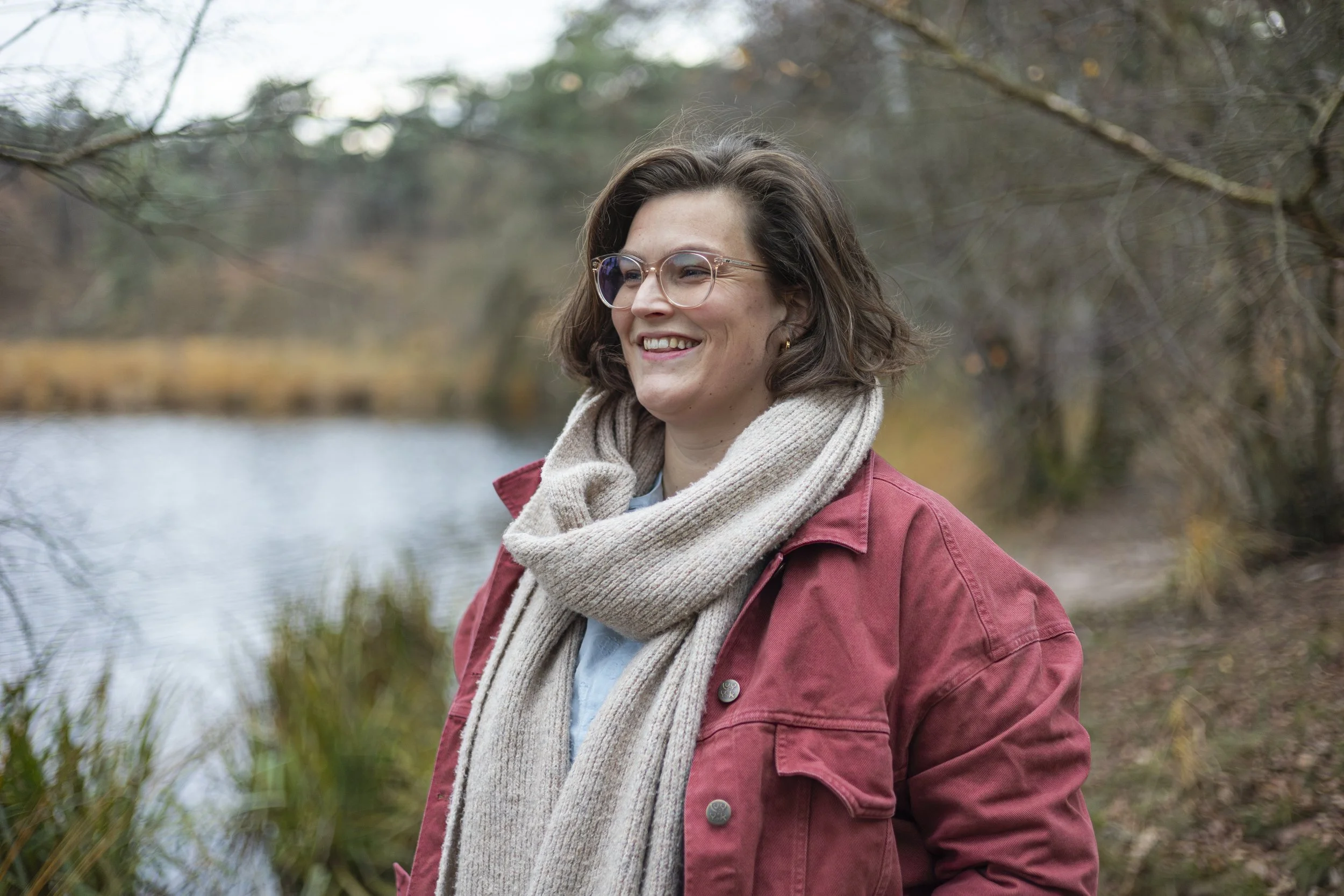 Vrouw met bril en rode jas wandelt langs meer in bosachtig gebied.