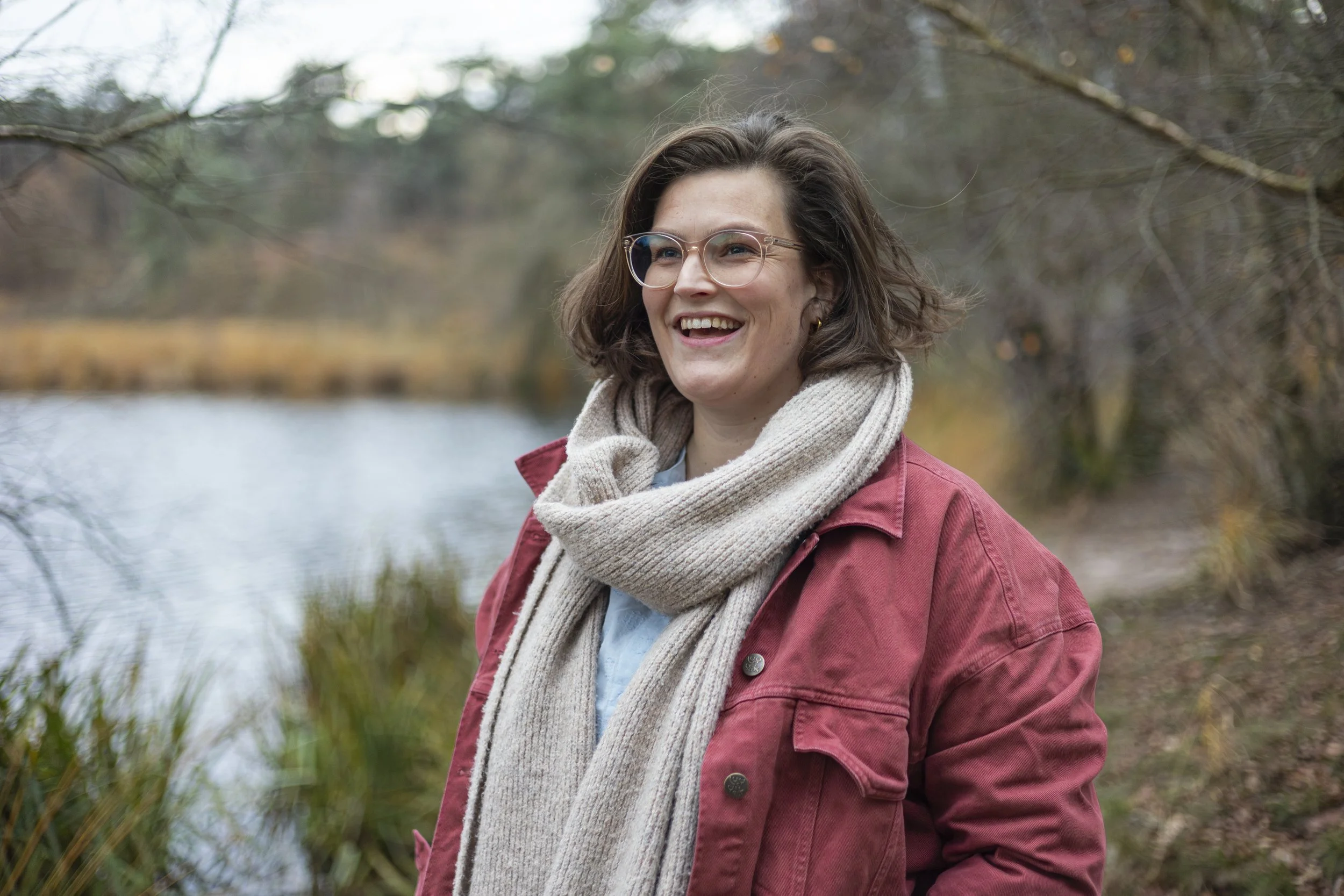 Vrouw met een bril en een rode jas staat buiten bij een meer, lacht en draagt een beige sjaal.