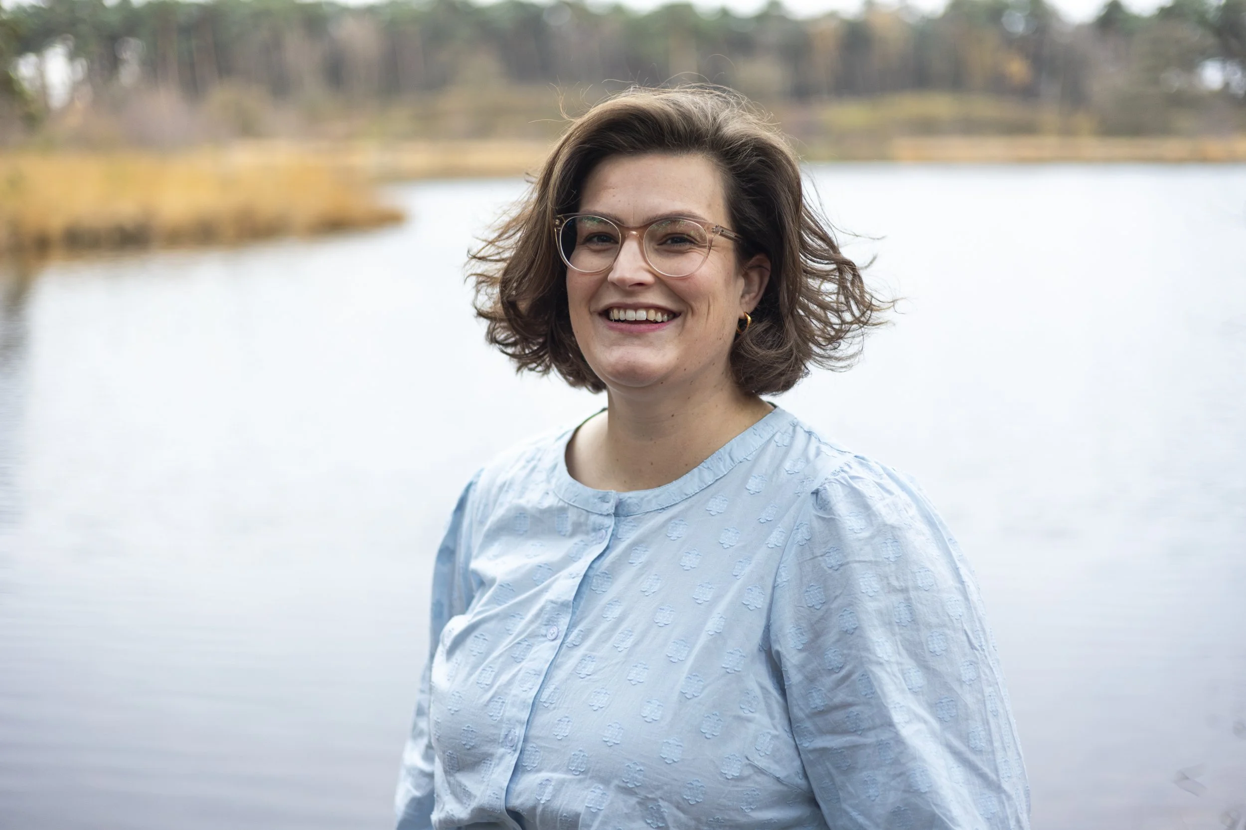 Vrouw met bril en blauw blouse staat lachend voor water en natuur op de achtergrond.