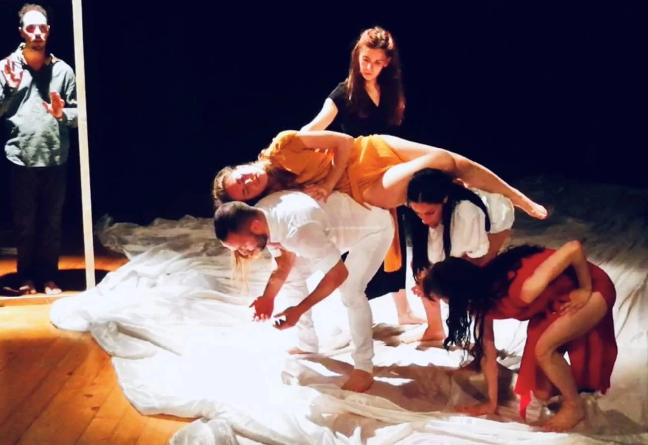 Teatro con attori che interpretano una scena con persone che si appoggiano l'una all'altra, alcune in ginocchio e altre in posizione seduta, con un attore in piedi a lato
