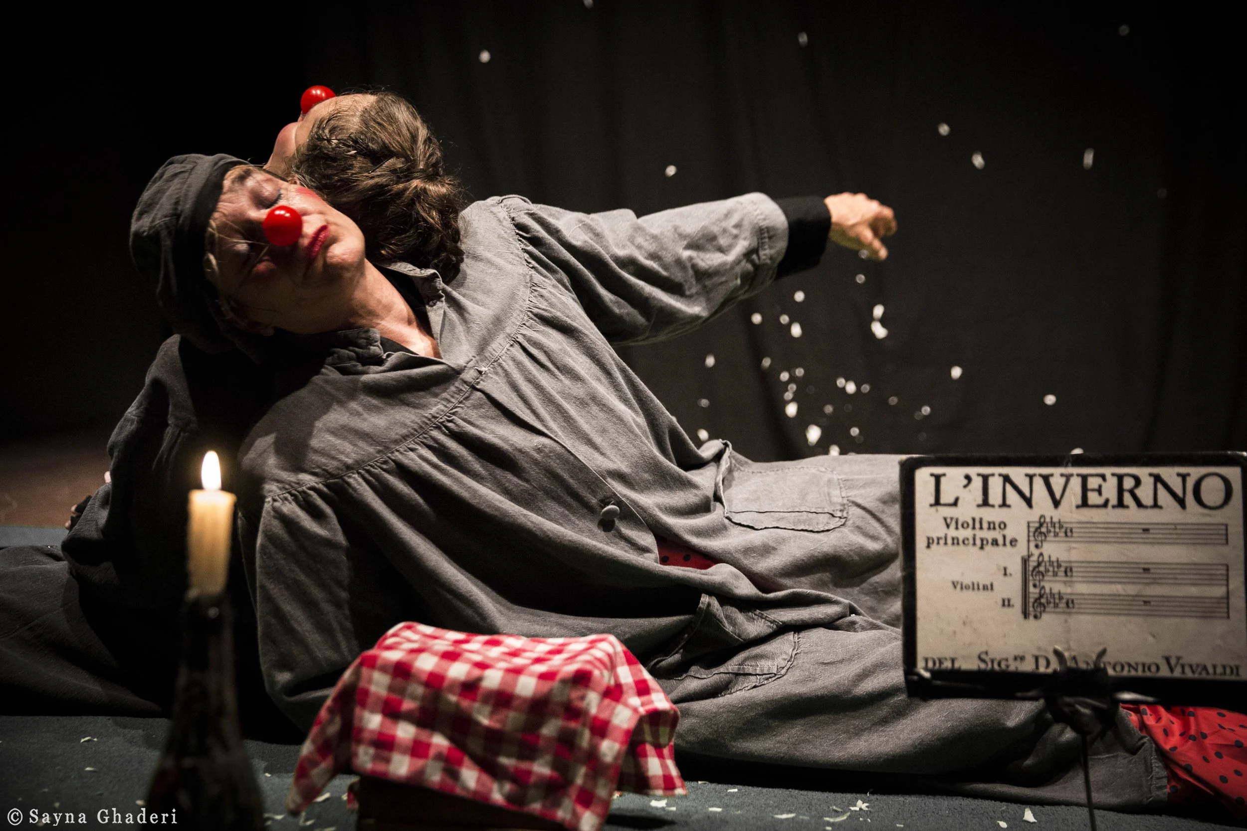 Una scena teatrale con due attrici vestite da clown con nasi rossi che simulano un incidente, una delle quali si è spostata indietro e l'altra si è piegata in avanti. Sul palco ci sono un candelabro con candela accesa e un cappello a scacchi rosso e bianco in primo piano. Un cartello indica "L'inverno" di Vivaldi e ci sono pezzi di carta che cadono dal soffitto.