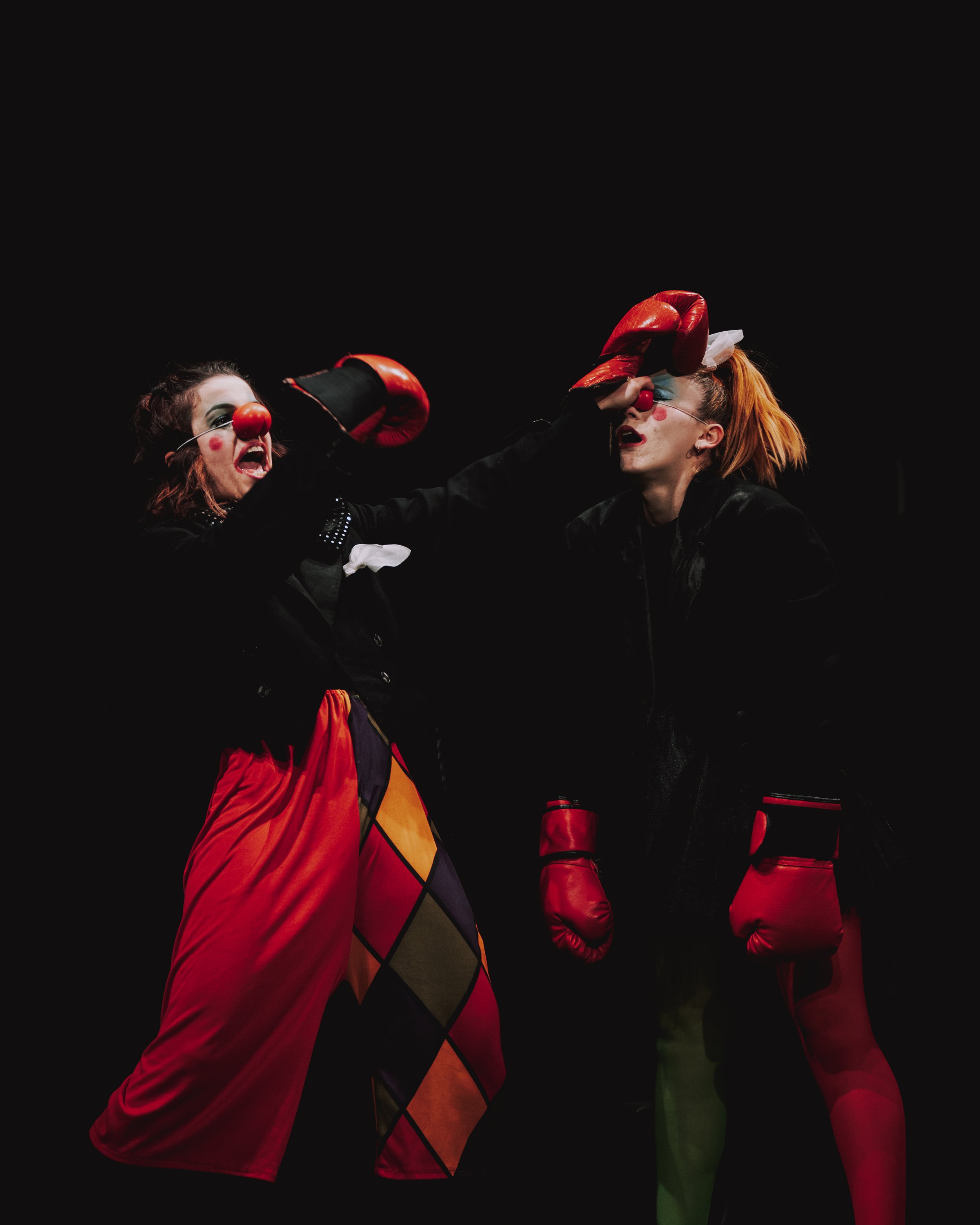 Una scena teatrale con due attrici vestite da clown con nasi rossi che simulano uno scontro di boxe