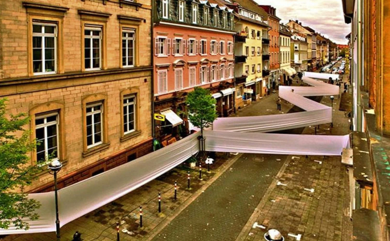 Strada nel centro storico di una città con edifici colorati, negozi e luci di lampioni, decorata con lunghe tende o drappi bianchi che si estendono lungo la via.