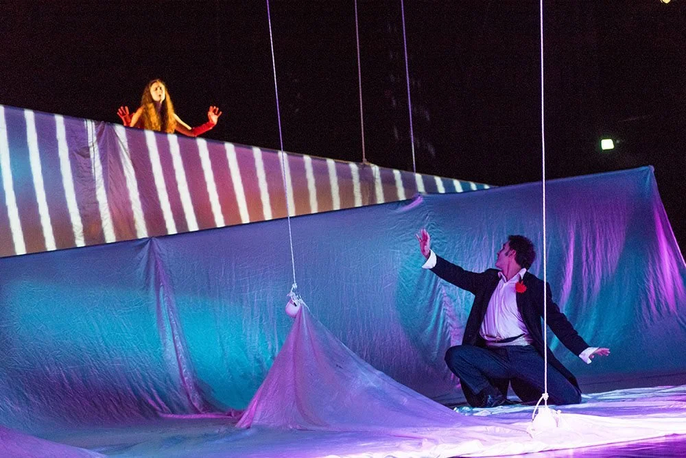 Una scena teatrale con due attori, uno in piedi sul palco e l'altro inginocchiato. La scena è illuminata con luci colorate e decorata con tessuto drappeggiato.