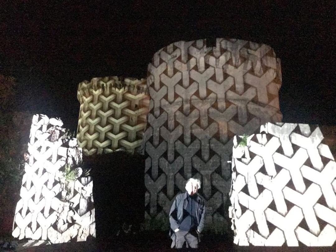 Persona con cappotto grigio davanti a sculture di grandi blocchi di pietra con motivi geometrici.