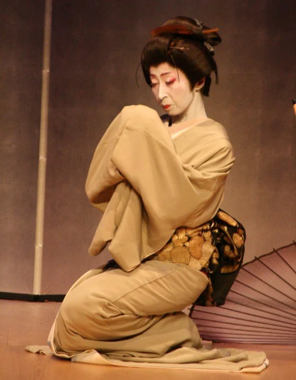 Una donna in abiti tradizionali giapponesi, con trucco e acconciatura classica, si inginocchia in una posa teatrale.