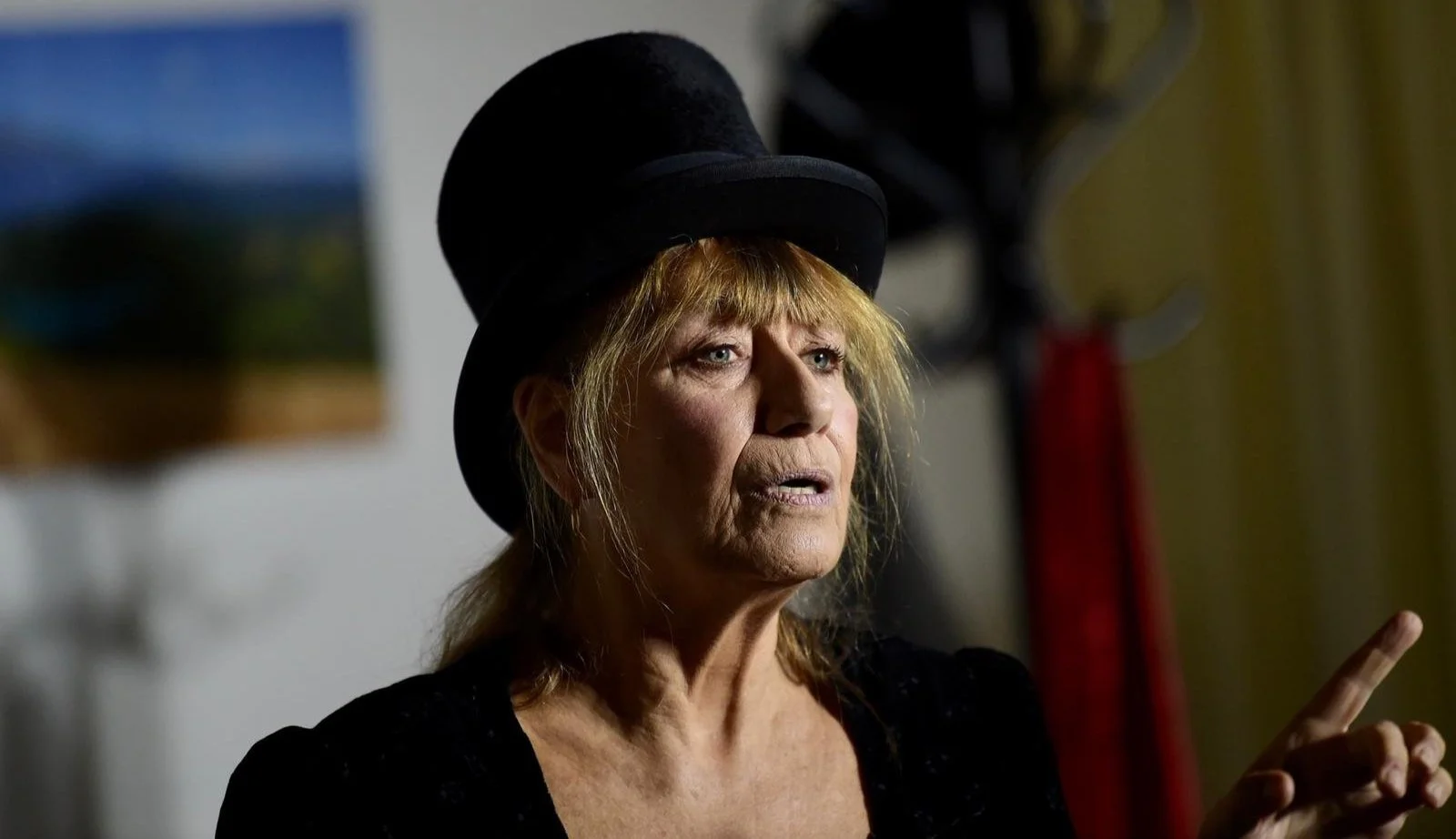 Donna anziana con cappello nero, capelli biondi, che parla o spiega con mano alzata.