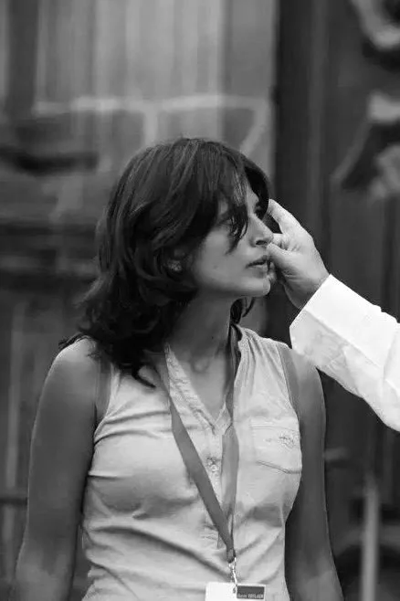 Donna con capelli corti e scuri, indossa una maglietta senza maniche e una tacca, mentre qualcuno le tocca il viso con la mano.