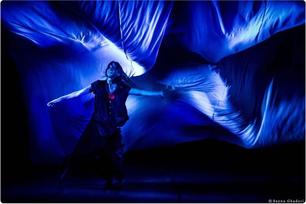 Una donna vestita con abiti scuri danza davanti a uno sfondo di tessuto blu illuminato, creando un effetto visivo artistico.