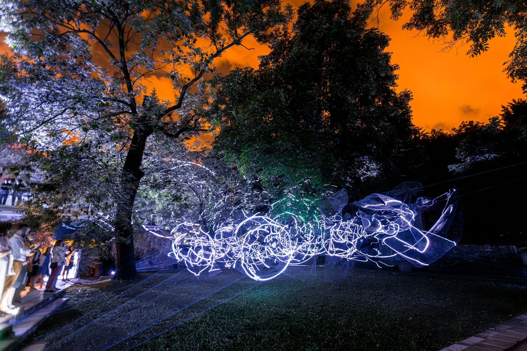 Un'installazione artistica luminosa in un parco con alberi durante il tramonto, con luci colorate che creano forme astratte sospese nell'aria, alcune persone si trovano sul lato sinistro dell'immagine.