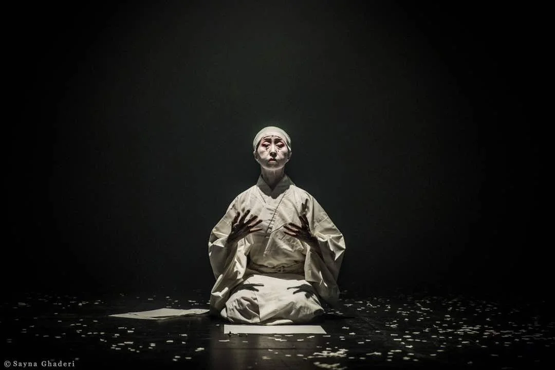 Una performer in costumi chiari si trova in posizione seduta sul palco, con un'espressione intensa, circondata da petali o foglie sparse, sotto una luce focalizzata in un ambiente scuro.