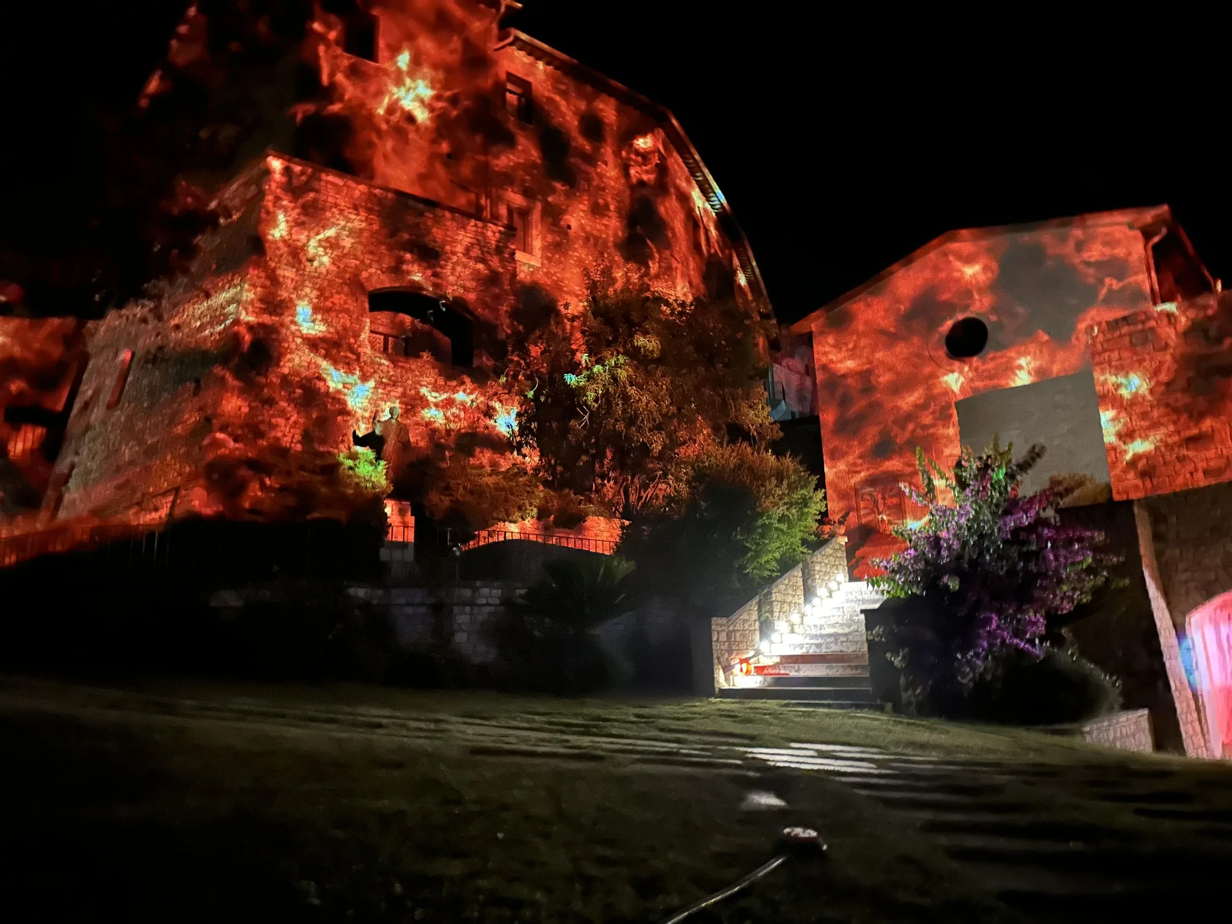 Impressionante scena notturna con un edificio che sembra bruciare, con projectioni di fiamme rosse sulle pareti e un giardino illuminato con fiori e luci.