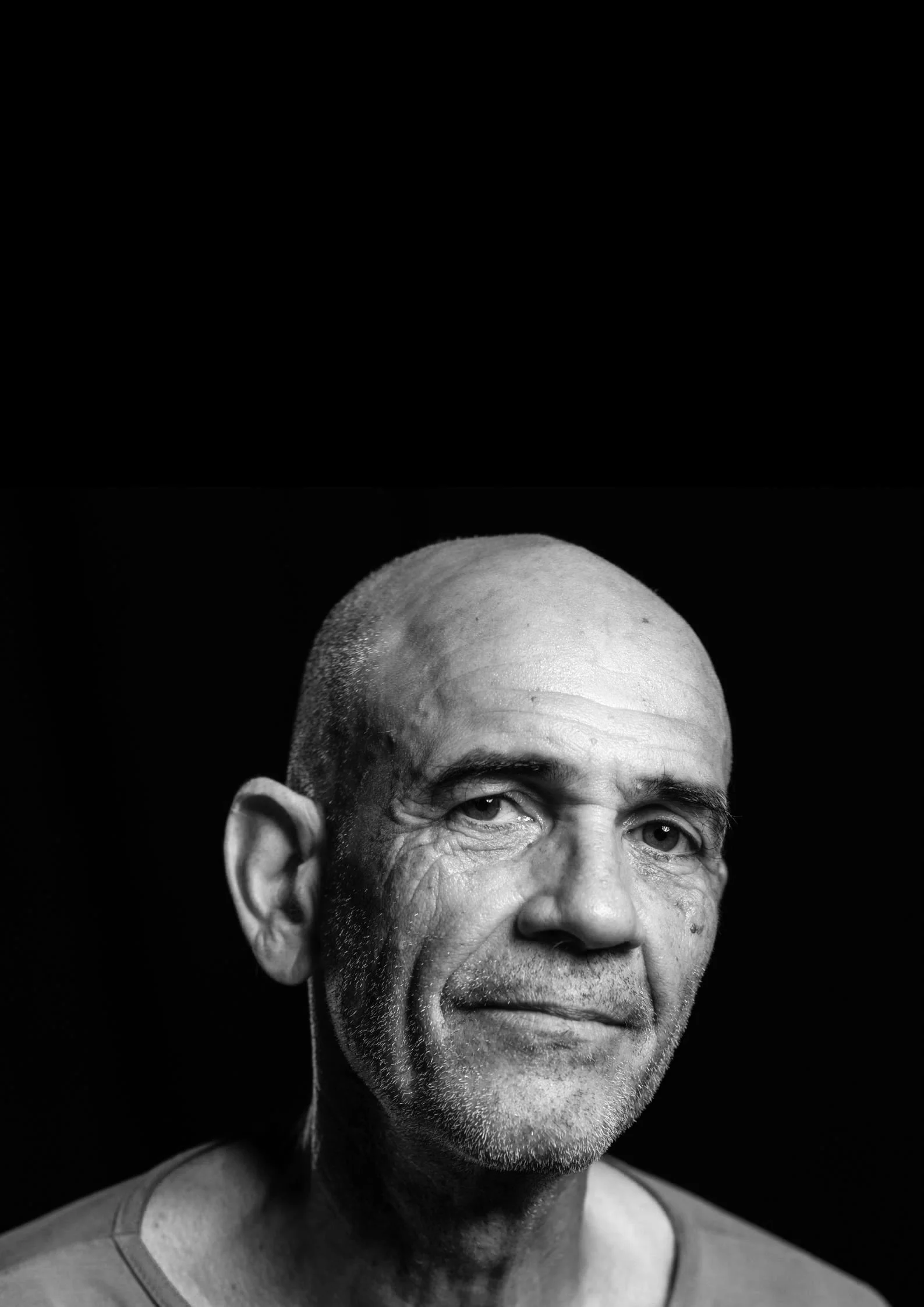 Ritratto in bianco e nero di un uomo anziano con pelle segnata, senza capelli, che indossa una maglietta. Lo sguardo è rivolto verso l'obiettivo, con un leggero sorriso.