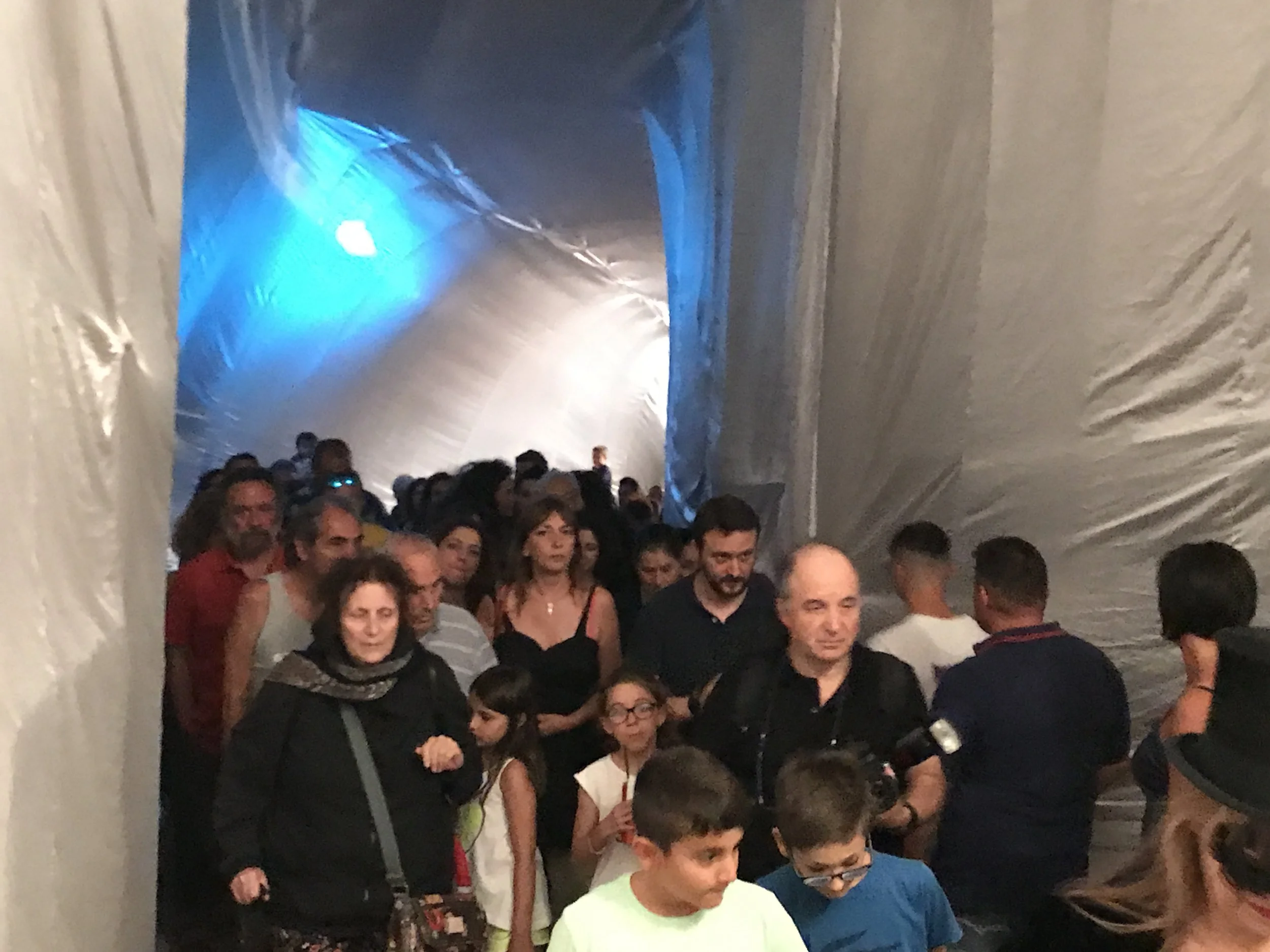 Gruppo di persone che camminano all'interno di un tunnel semi-chiuso, illuminato da luci blu e bianche, con pareti di plastica o tessuto.