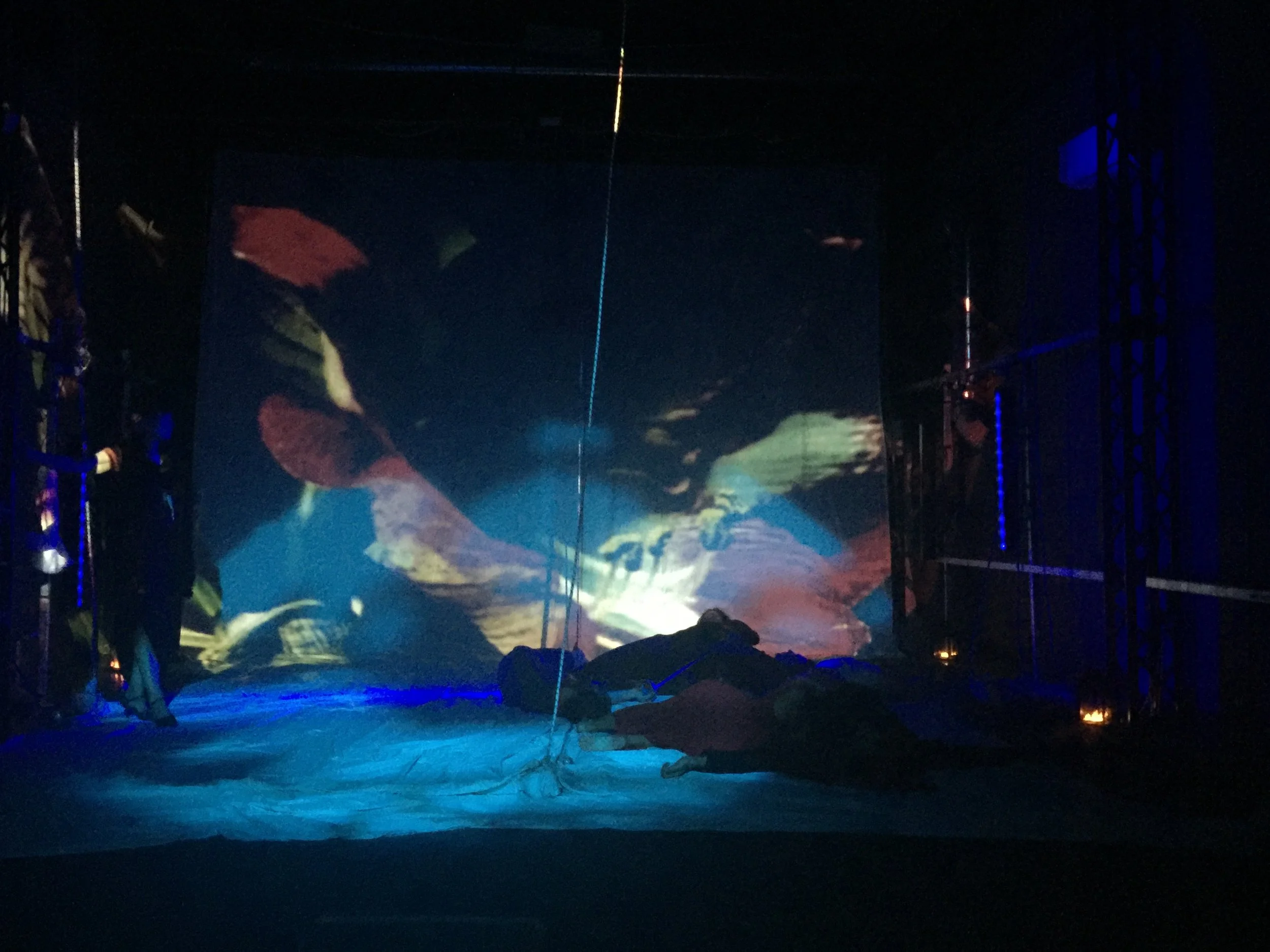 Una scena teatrale con persone sdraiate sul palco davanti a uno schermo che proietta immagini astratte e colorate, con illuminazione soffusa e luci blu.