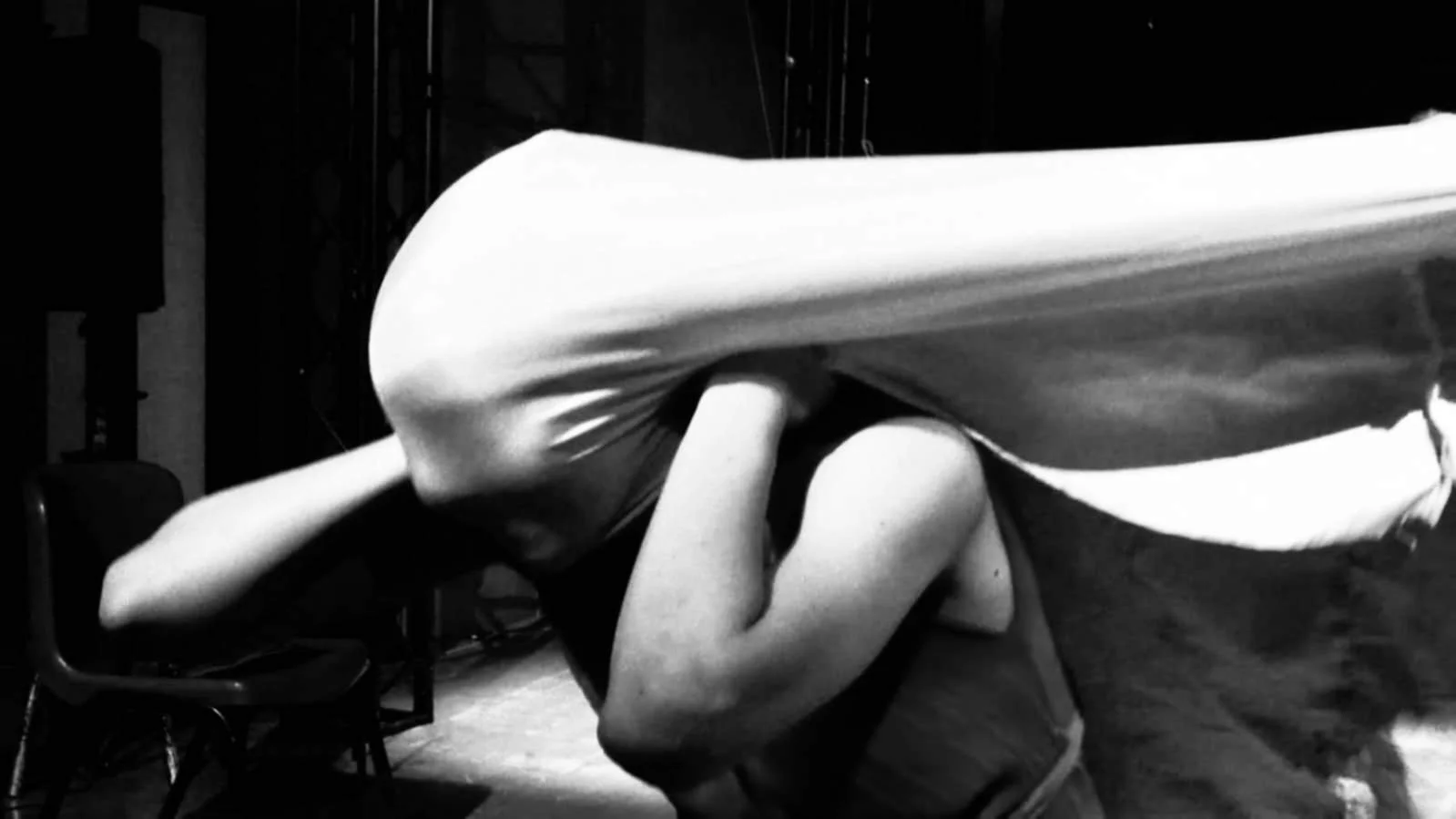 Figura umana distesa a terra con il volto nascosto, avvolta da un tessuto o tenda, in un ambiente interno con sedie intorno.