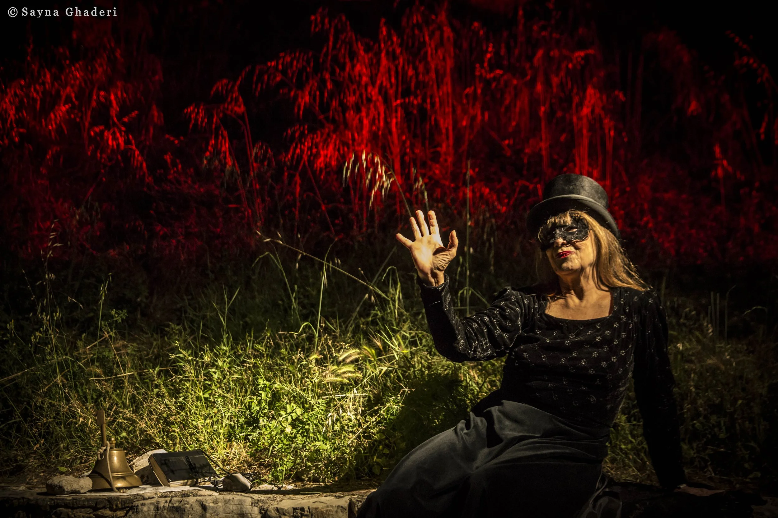 Una donna con cappello nero e maschera nera seduta a terra, al centro dell'immagine, con una mano alzata, in un ambiente esterno con vegetazione, illuminata da luci calde e uno sfondo rosso intenso.