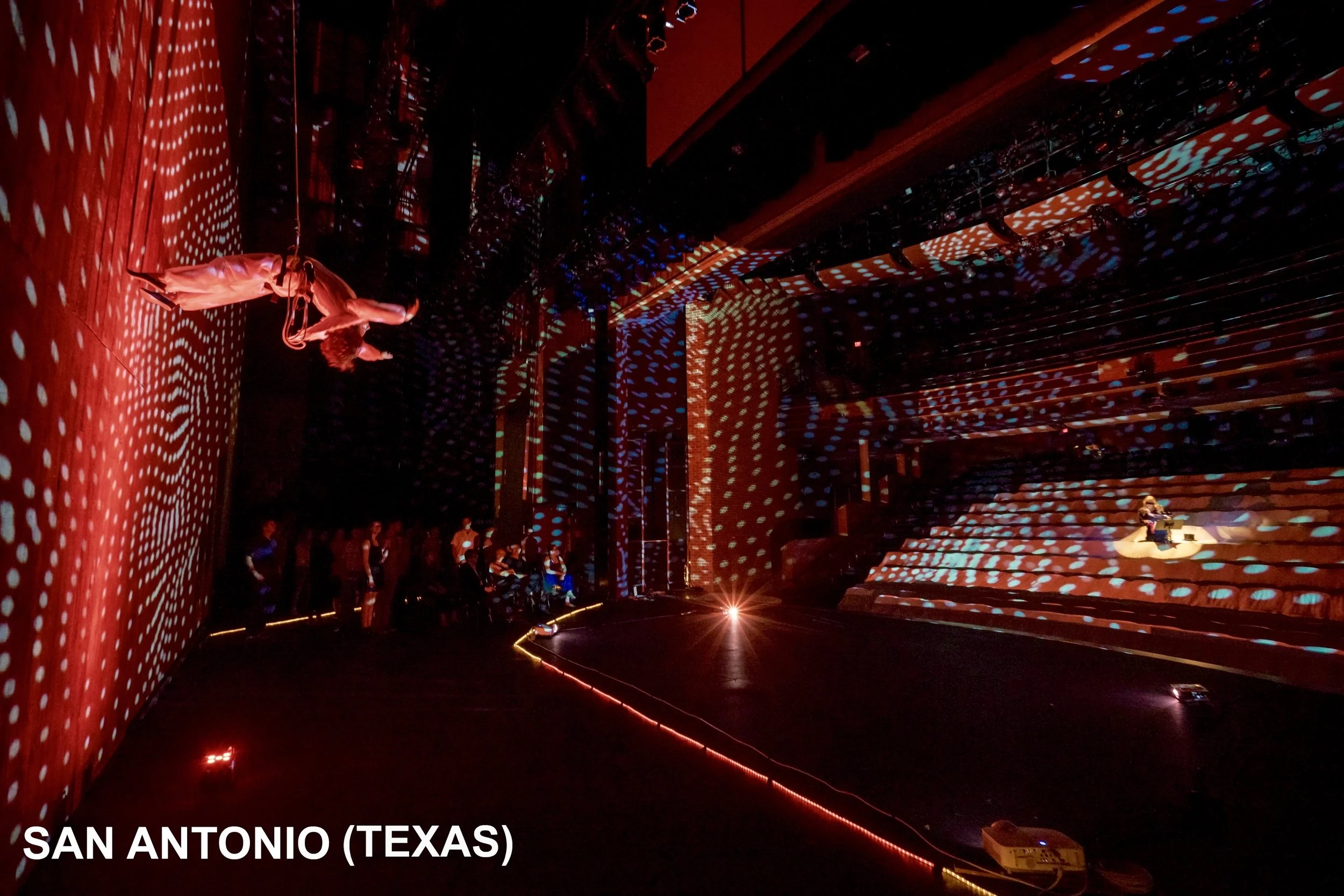 Interno di un teatro con luci rosse e blu, con pareti e gradinate illuminate con motivi di punti bianchi e blu, sul palco si vedono due performer, una di fronte e una a destra su una sedia, con antifurto e con pubblico in ombra. In basso a sinistra, 