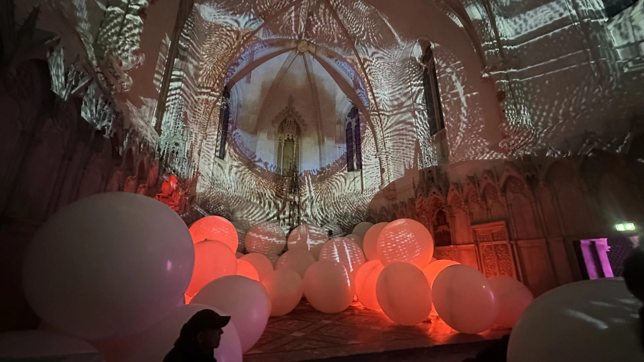 Interno di una chiesa con proiezioni di luci e pattern sulla volta e pareti, e grandi sfere di luce rossa e bianca sul pavimento.