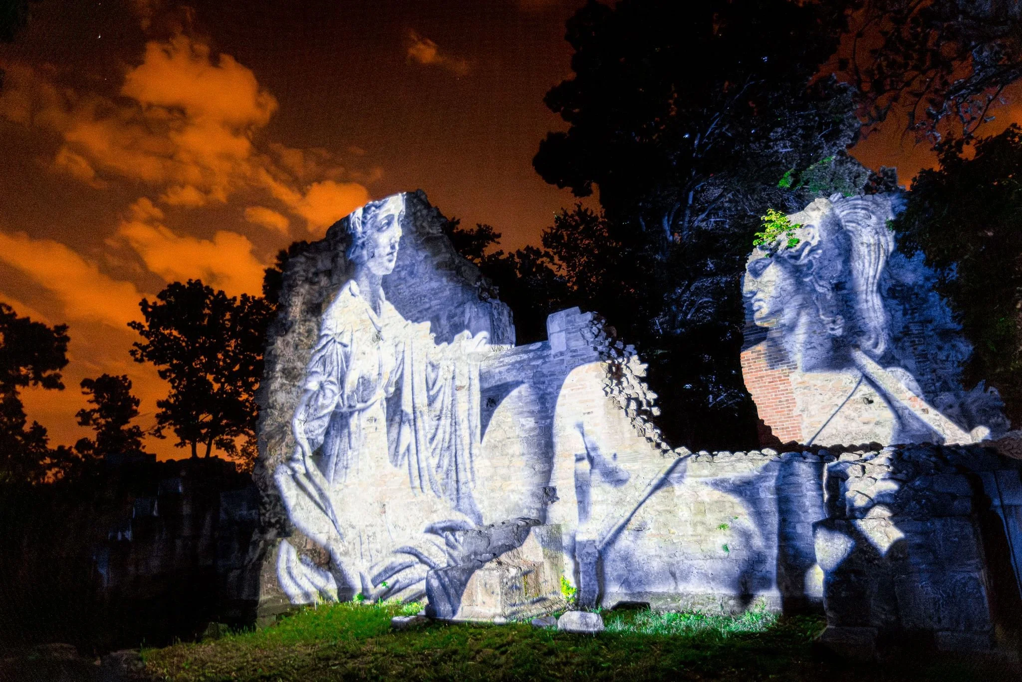 Scultura lignea illuminata di notte con sfondo di cielo arancione e nuvole, circondata da alberi.