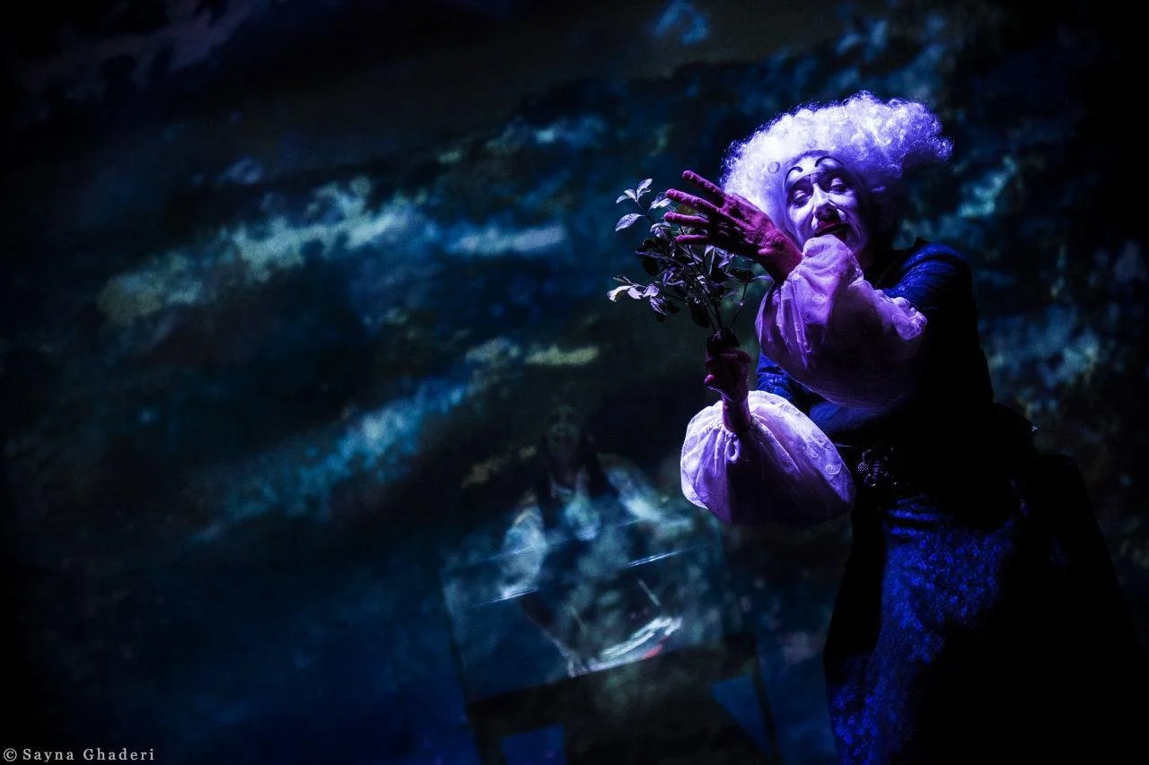 Una figura teatrale vestita in modo elaborato con trucco e parrucca bianca, che posa con una foglia in mano, in un'illuminazione bluoscura.