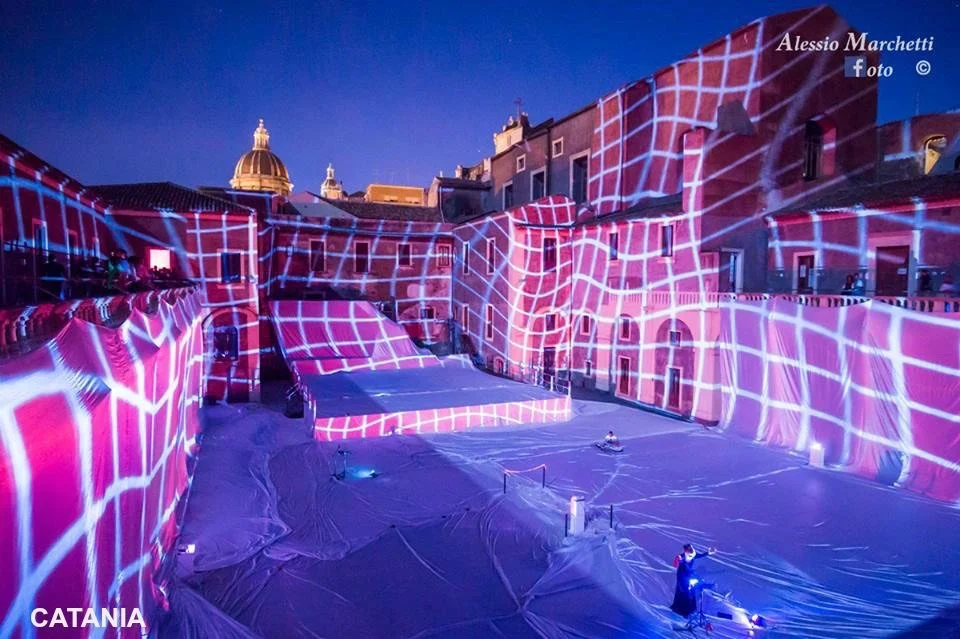 Una scena urbana con edifici antichi illuminati da proiezioni luminose a scacchi di colore rosa e blu, durante la notte, a Catania.