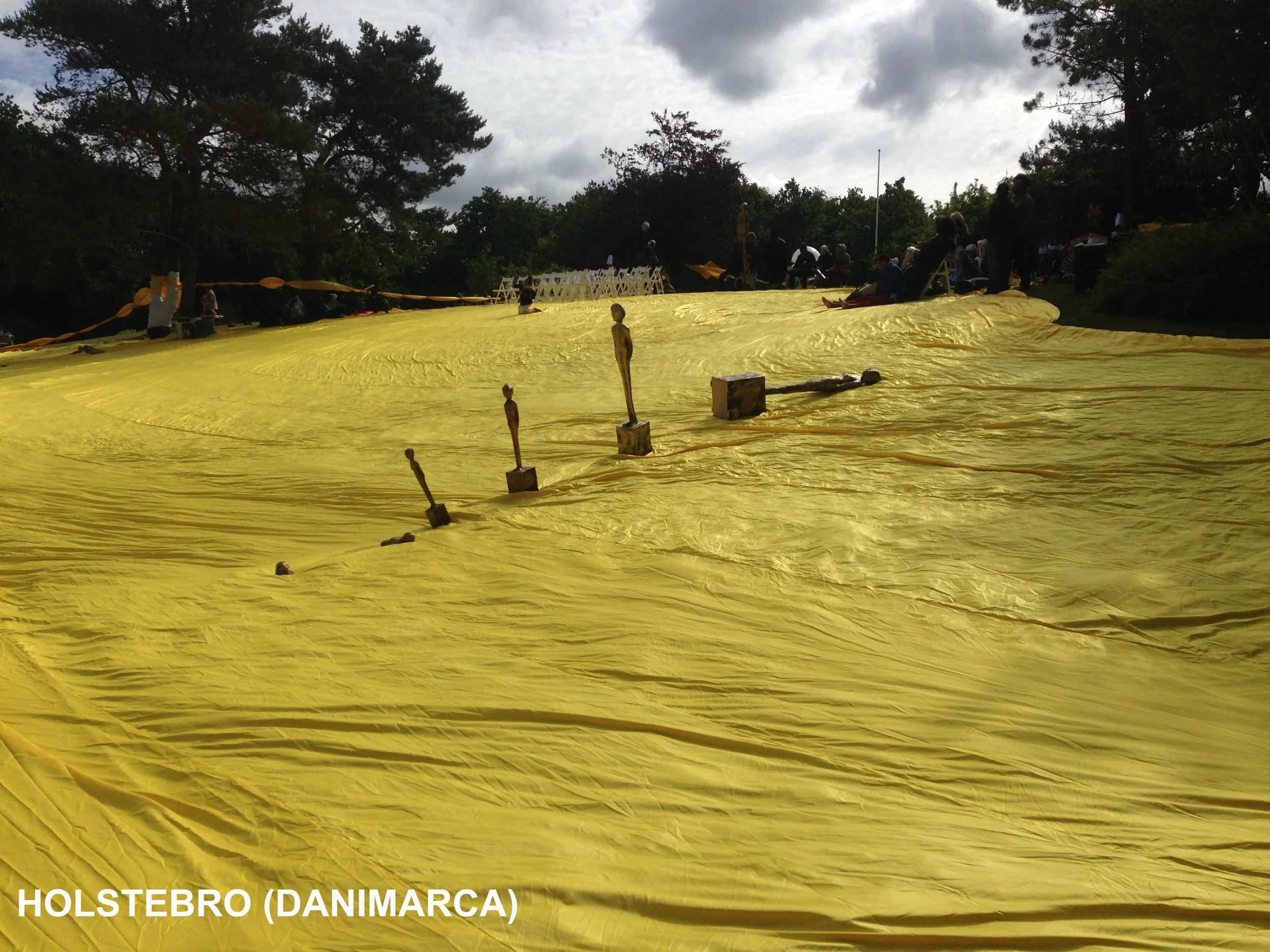 Una grande tela gialla stesa su un prato, con alcune piccole sculture di figure umane disposte sopra. In lontananza, persone sedute sui bordi e in piedi, sotto un cielo nuvoloso, vicino a degli alberi. L'immagine è stata scattata a Holstebro, Danimar