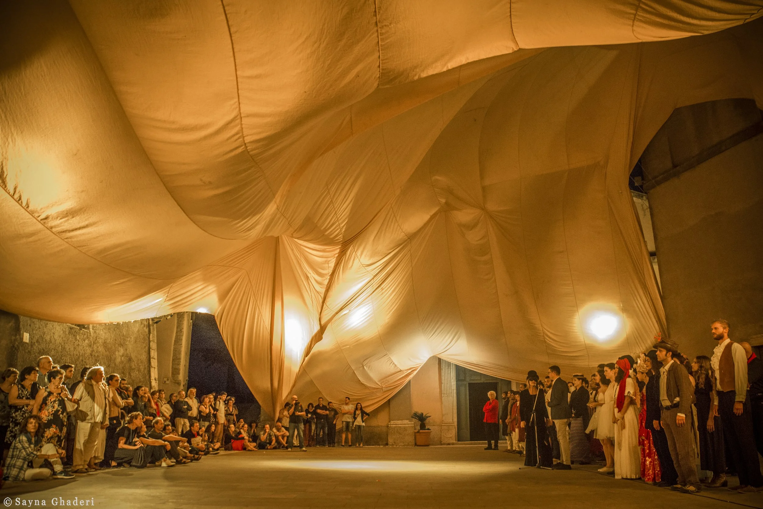 Un gruppo di persone con abiti cavallereschi e vestiti storici si riunisce in un'ampia sala con un soffitto ingegneristico dorato. Alcuni sono seduti a terra, altri in piedi, mentre assistono a uno spettacolo o a un evento culturale.