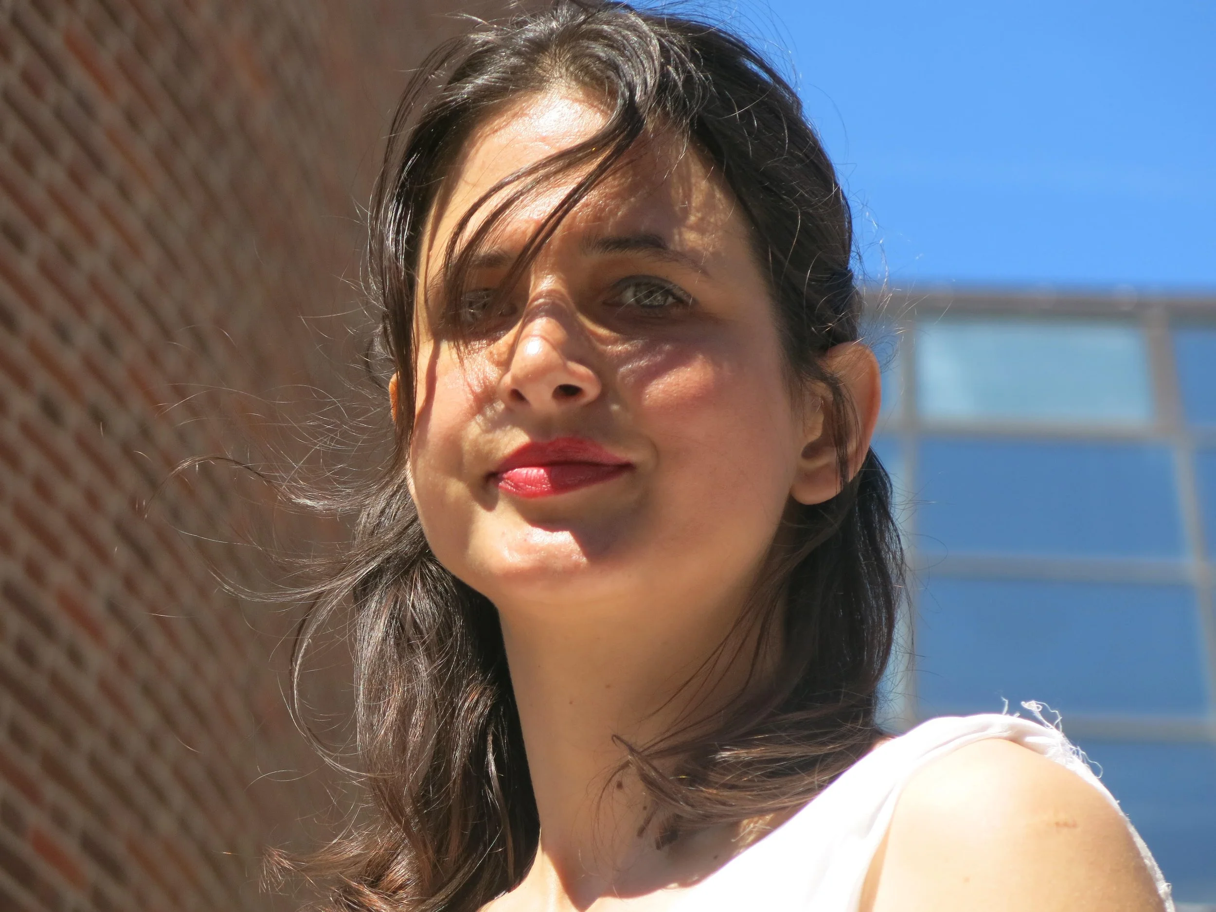 Ragazza con capelli castani, sorriso, in un ambiente urbano, con un muro di mattoni a sinistra e un edificio e il cielo blu sullo sfondo.