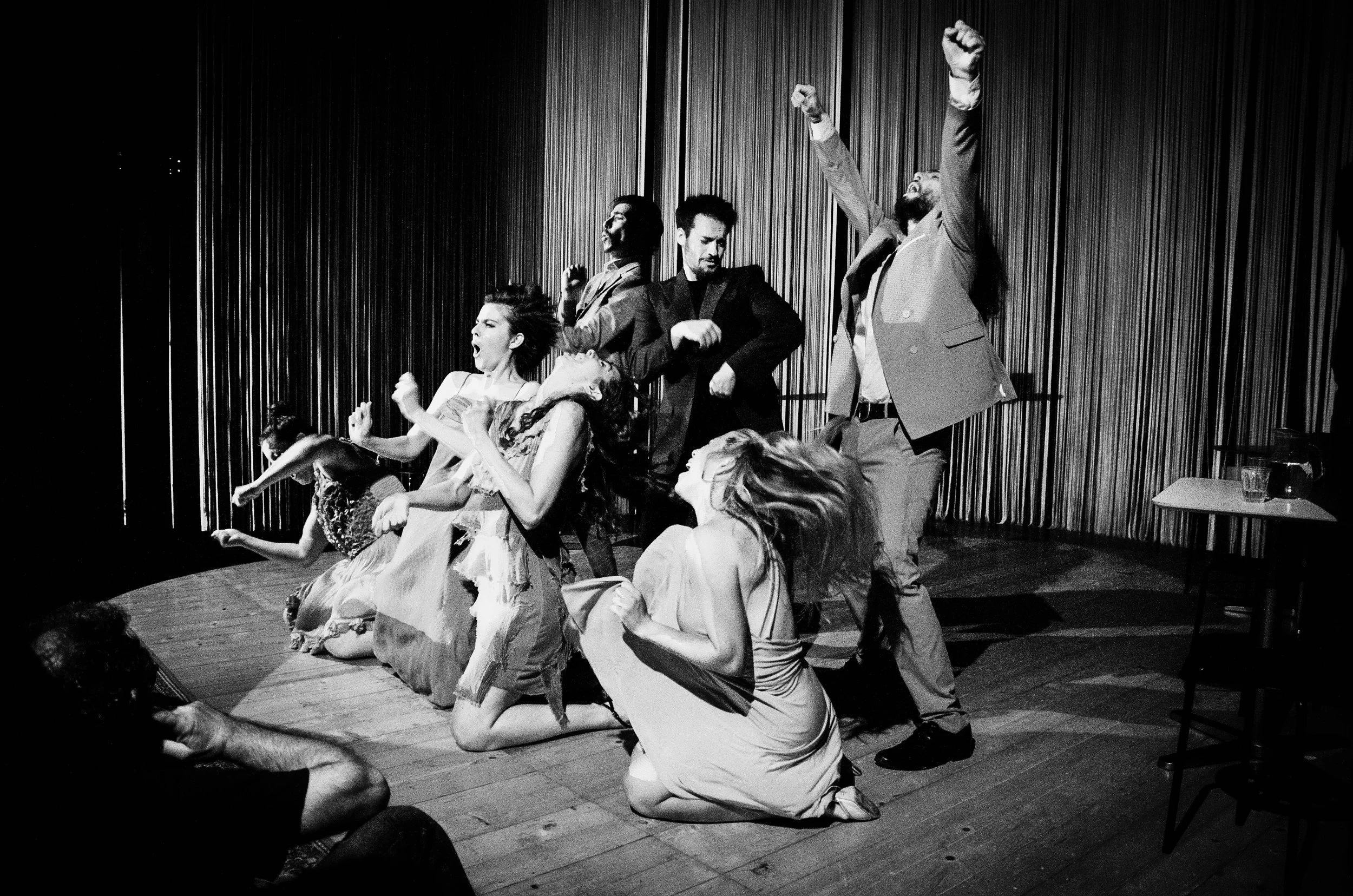 Gruppo di persone che recitano o ballano su un palco con tende geometriche sullo sfondo, alcune sono in piedi, altre sono inginocchiate o sedute, con espressioni intense e movimenti dinamici.