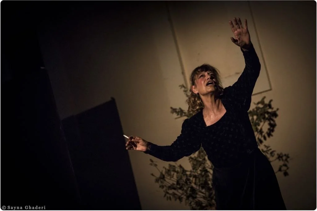 Donna con abbigliamento nero e capelli riccioli, che si muove con un’espressione emotiva, con una mano alzata e l’altra che tiene un oggetto sottile, probabilmente una sigaretta, in un ambiente interno con piante sullo sfondo.