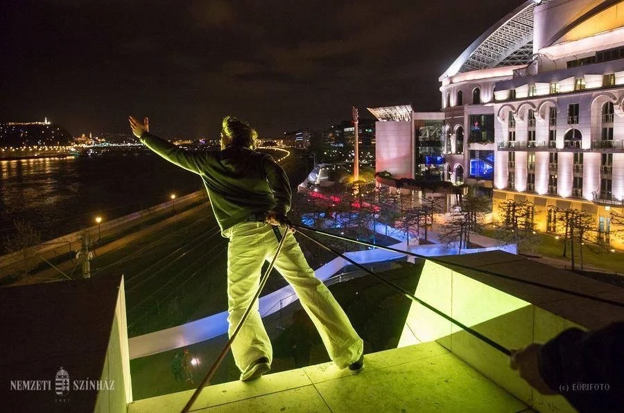 Persona su una grondaia di notte davanti a un edificio illuminato con luci colorate.