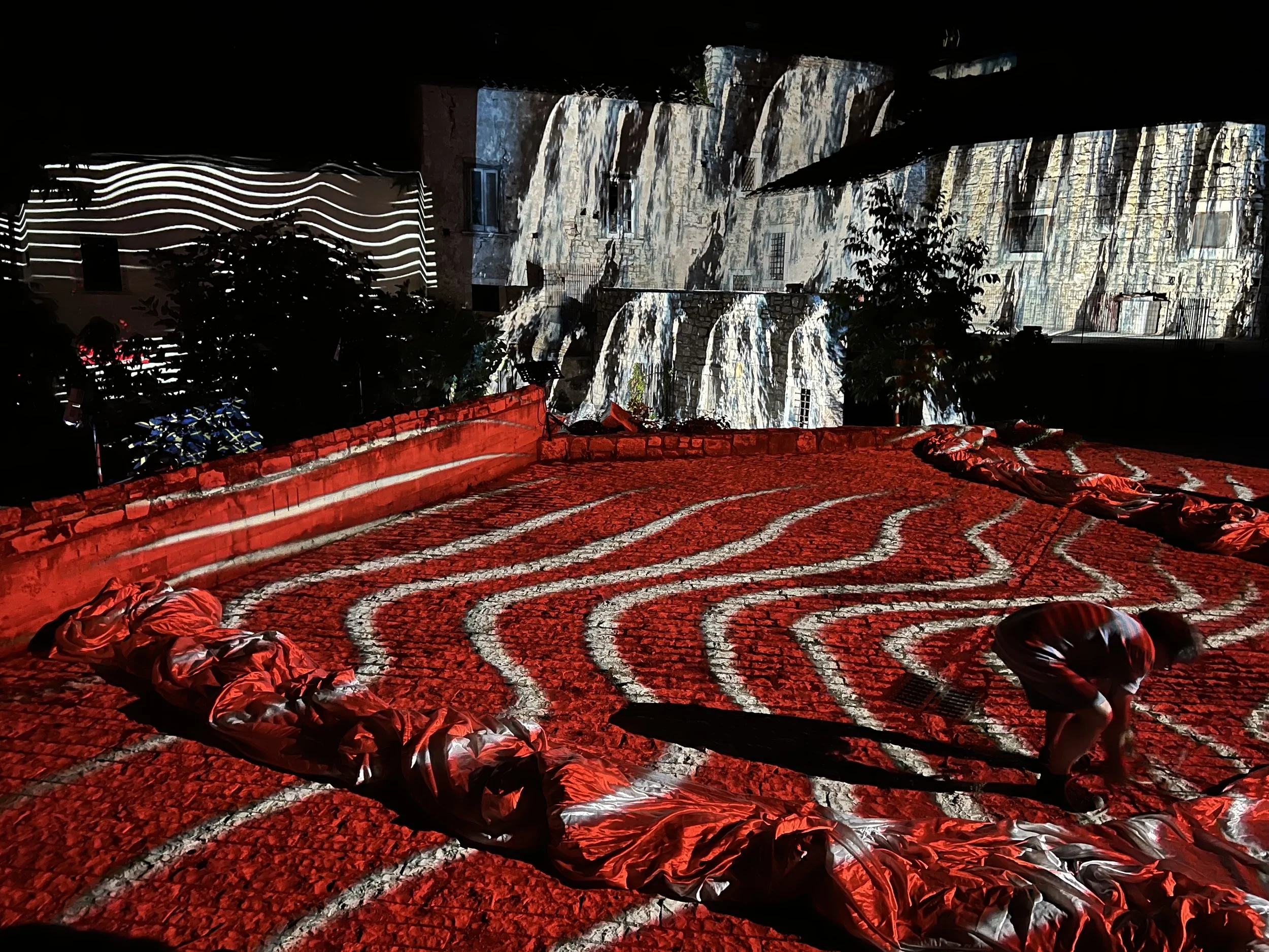 Installazione artistica all'aperto con luci e proiezioni di righe luminose su una superficie rossa e su edifici vicini, con una persona che lavora sul pavimento.