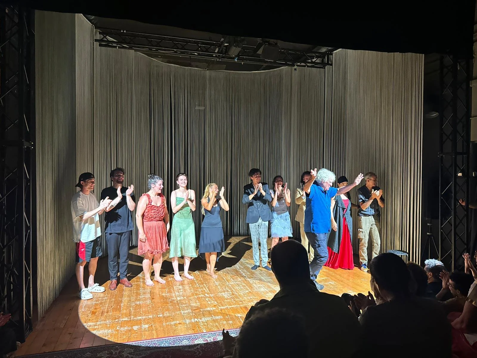 Un gruppo di persone saluta sul palco di uno spettacolo, con alcuni che applausano e altri che si inchinano, sotto una tenda con sfondo di tendaggi dorati.