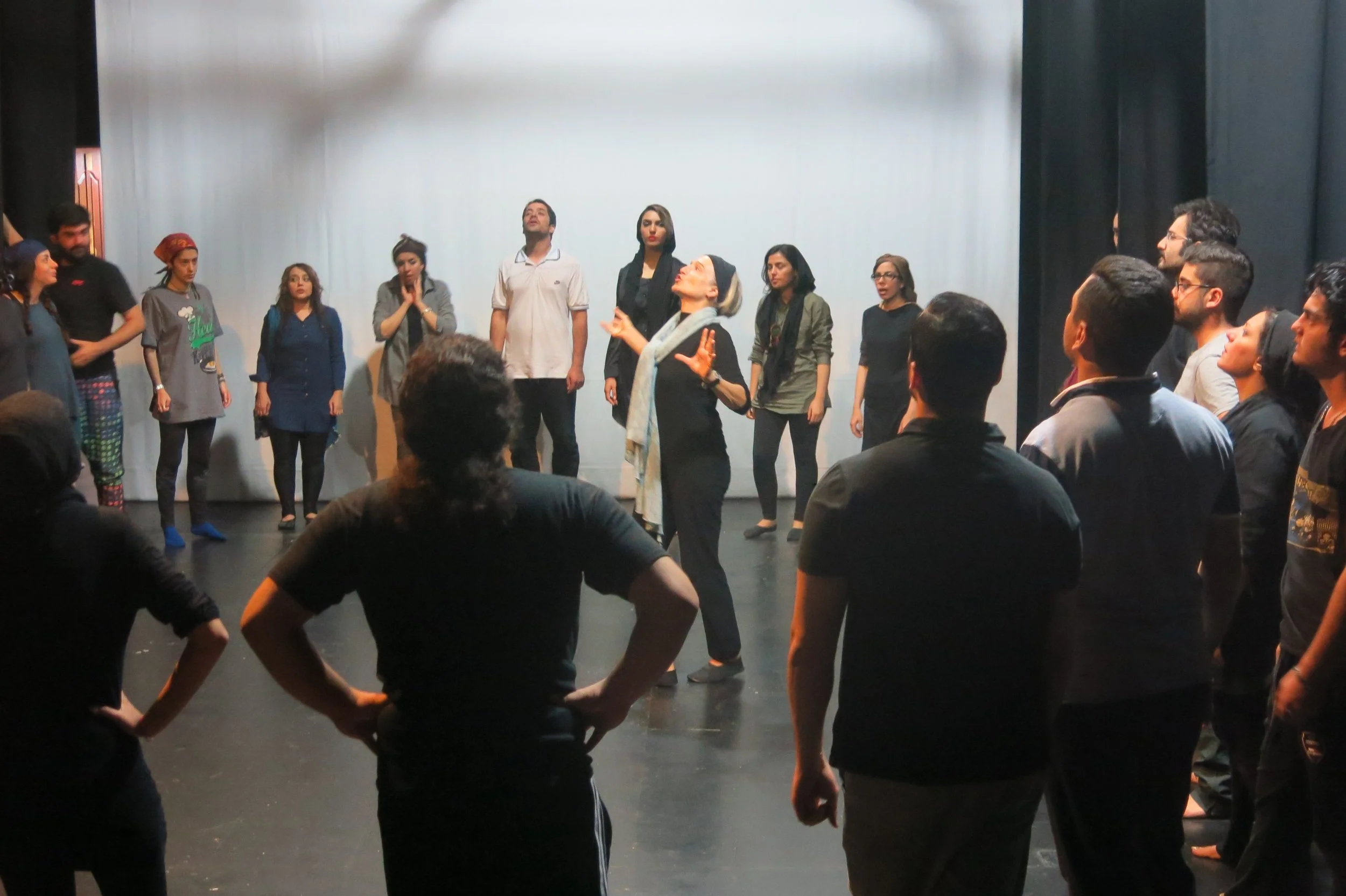Un gruppo di persone si trova in uno studio teatrale, con una donna al centro che sembra dirigere o spiegare una scena, circondata da altri attori o partecipanti con varie espressioni e posizioni.