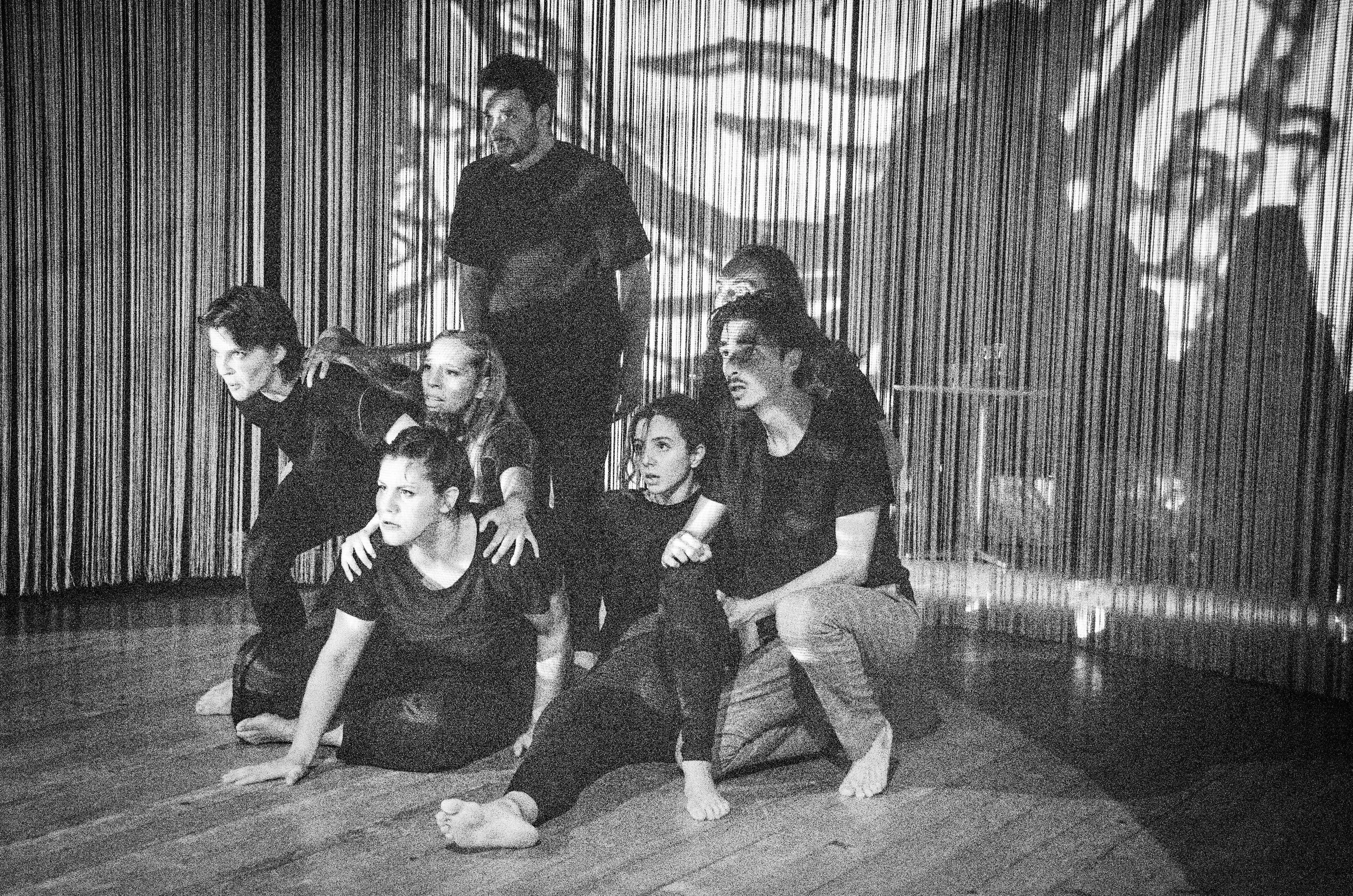 Gruppo di sette persone in pose drammatiche, alcune sedute e altre inginocchiate, in uno spazio con sfondo di tende a righe. L'immagine è in bianco e nero.