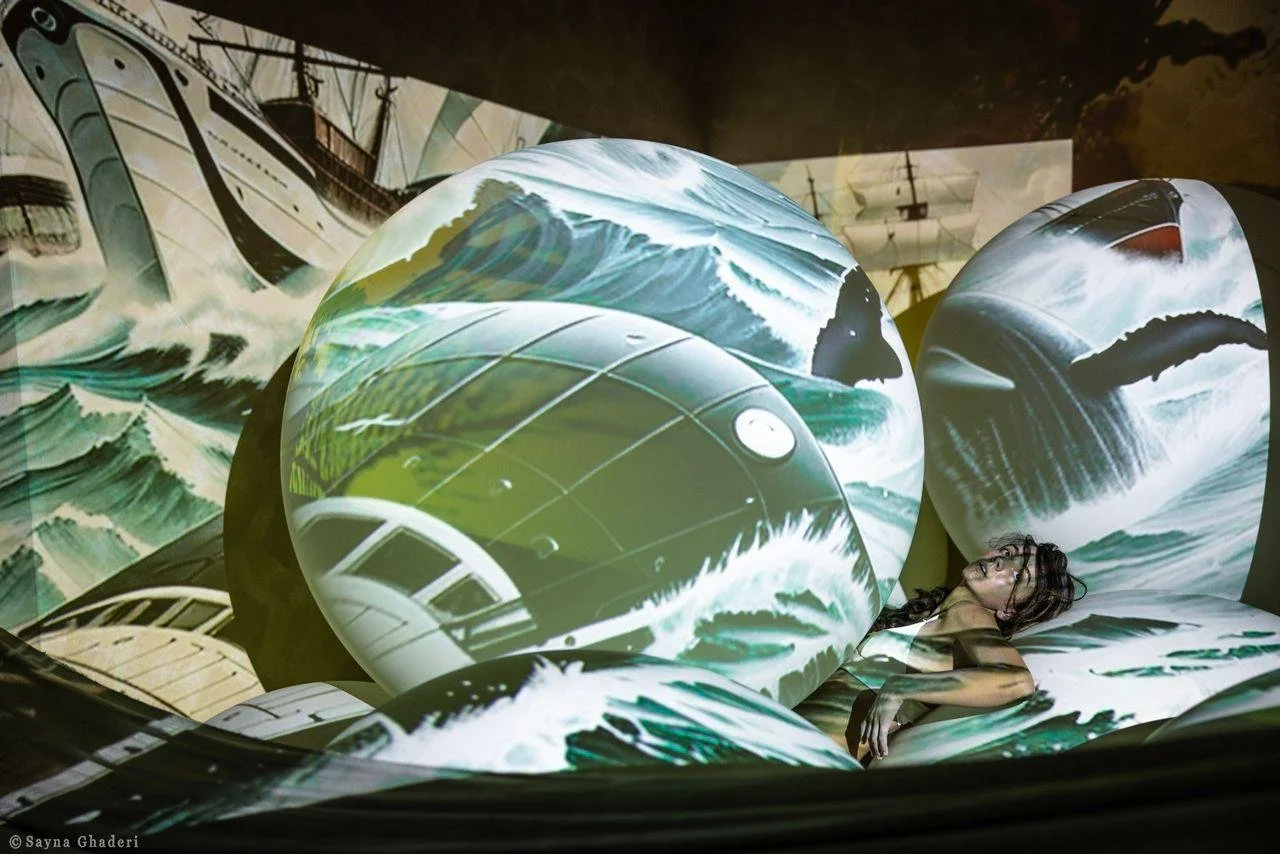mostra immersiva con immagini di onde e navi proiettate su sfere e pareti, con donna distesa tra le proiezioni