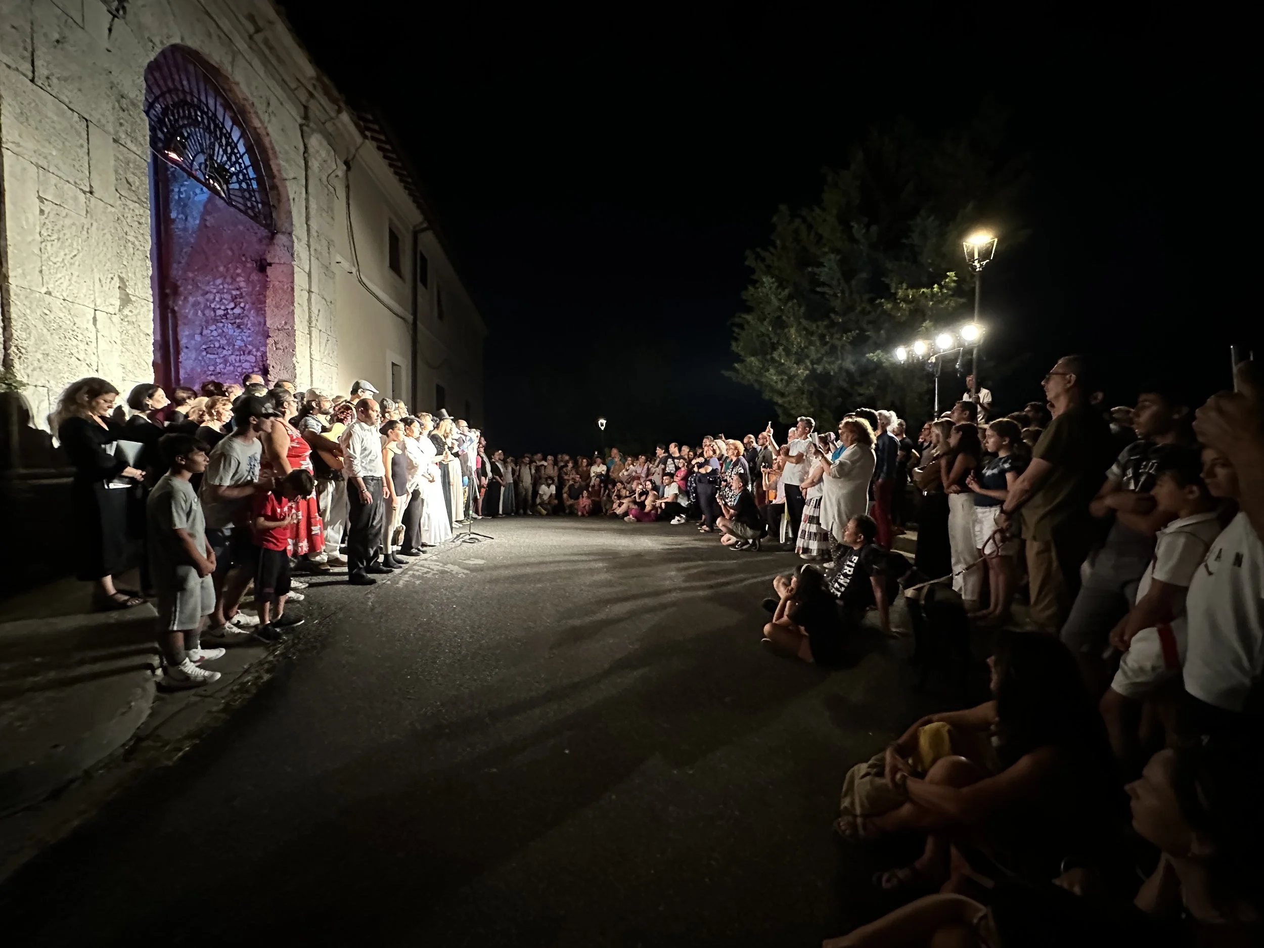 Gruppo di persone di diverse età che si riuniscono all'aperto di notte davanti a un edificio con arca e illuminazione. La scena è festosa e pubblica.