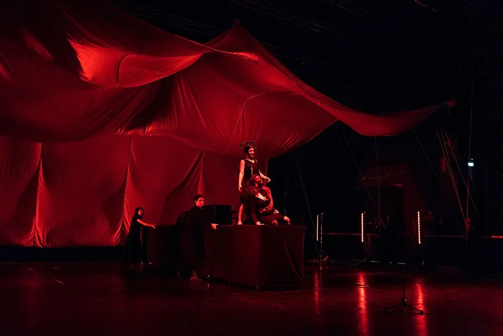 Persone su un palcoscenico con una grande drappo rosso come sfondo, sembianze di un'esibizione teatrale o di un concerto, illuminazione rossa.