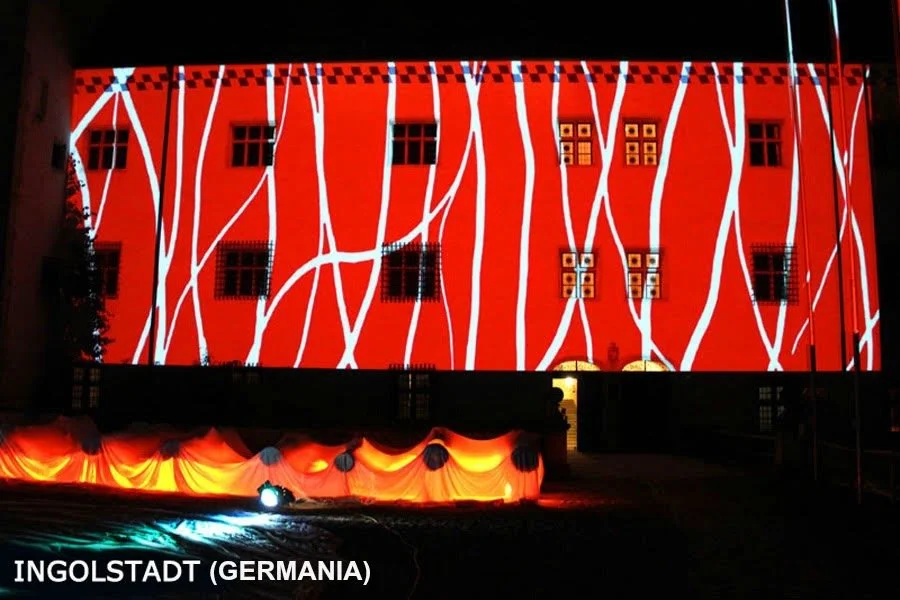 Proiezione di luci e disegni sul lato di un edificio rosso a Ingolstadt, Germania, con linee bianche che attraversano la facciata.