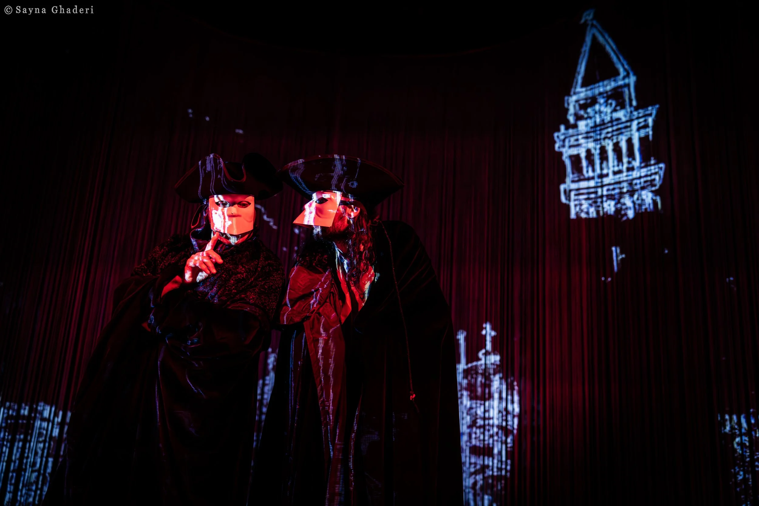 Due figure mascherate con abiti oscuri e cappelli tricorno, indossano maschere che coprono il volto, in un ambiente con sfondo rosso scuro e proiezioni luminose di edifici storici.