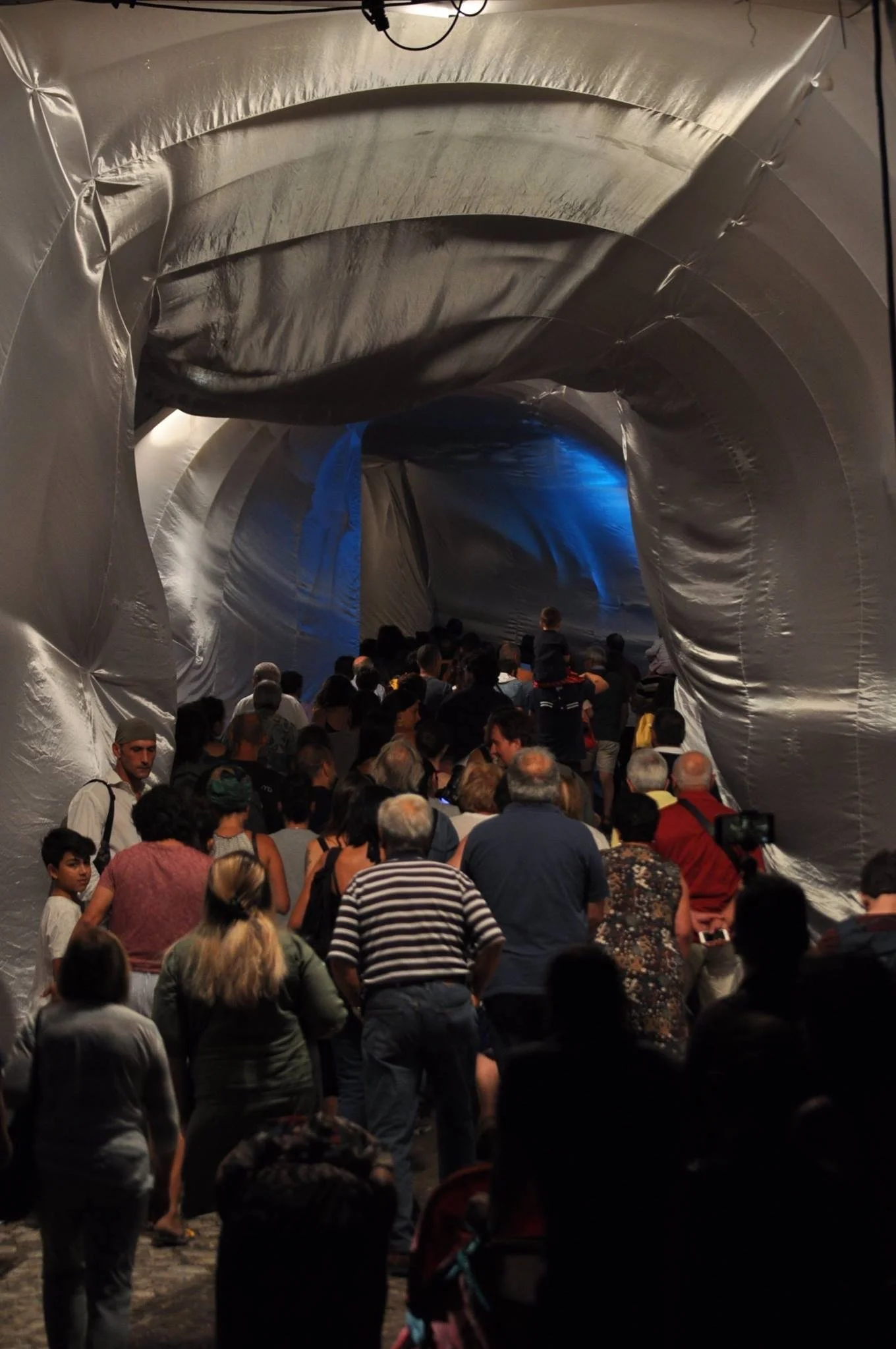 Folla di persone in fila in un tunnel di materiali metallici, sembra un evento pubblico o un'installazione artistica.