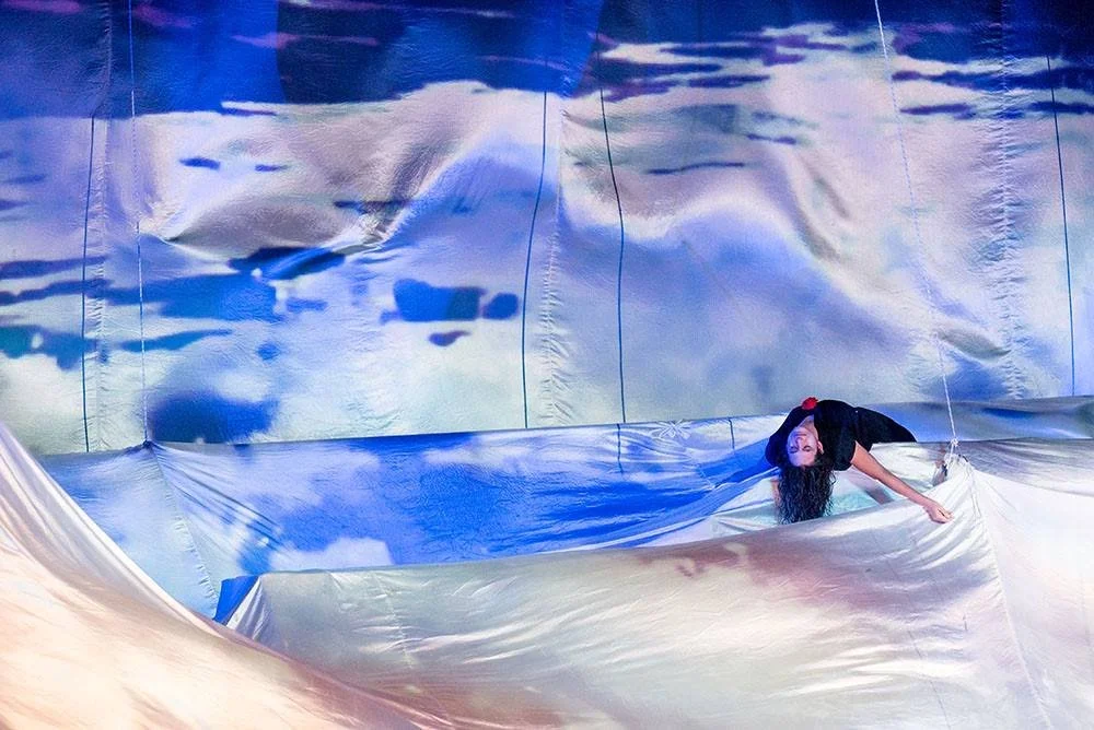 Una donna si trova su una scena teatrale con sfondo e superfici di tessuto colorato, che simula un cielo, creando un'illusione di nuvole e cielo.