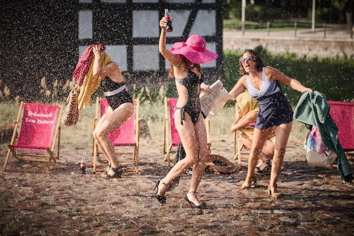 Un gruppo di donne si diverte sulla spiaggia sotto la pioggia, indossando costumi da bagno, cappelli grandi, e occhiali da sole, mentre ridono e giocano.