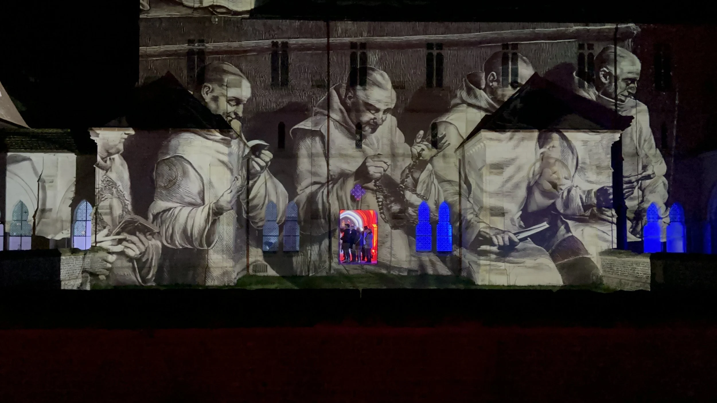 Proiezione di un'illustrazione in bianco e nero di persone che leggono e scrivono sui muri di un edificio storico, con finestre di vetro colorate blu e rossa e una porta di ingresso con luci rosse.