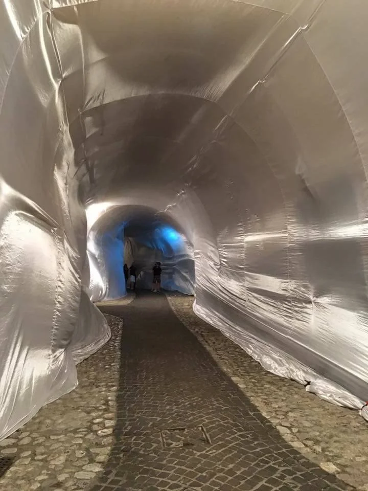 Tubo di metallo rivestito di un materiale riflettente, con una strada acciottolata che passa al suo interno, e alcune persone che camminano in fondo.